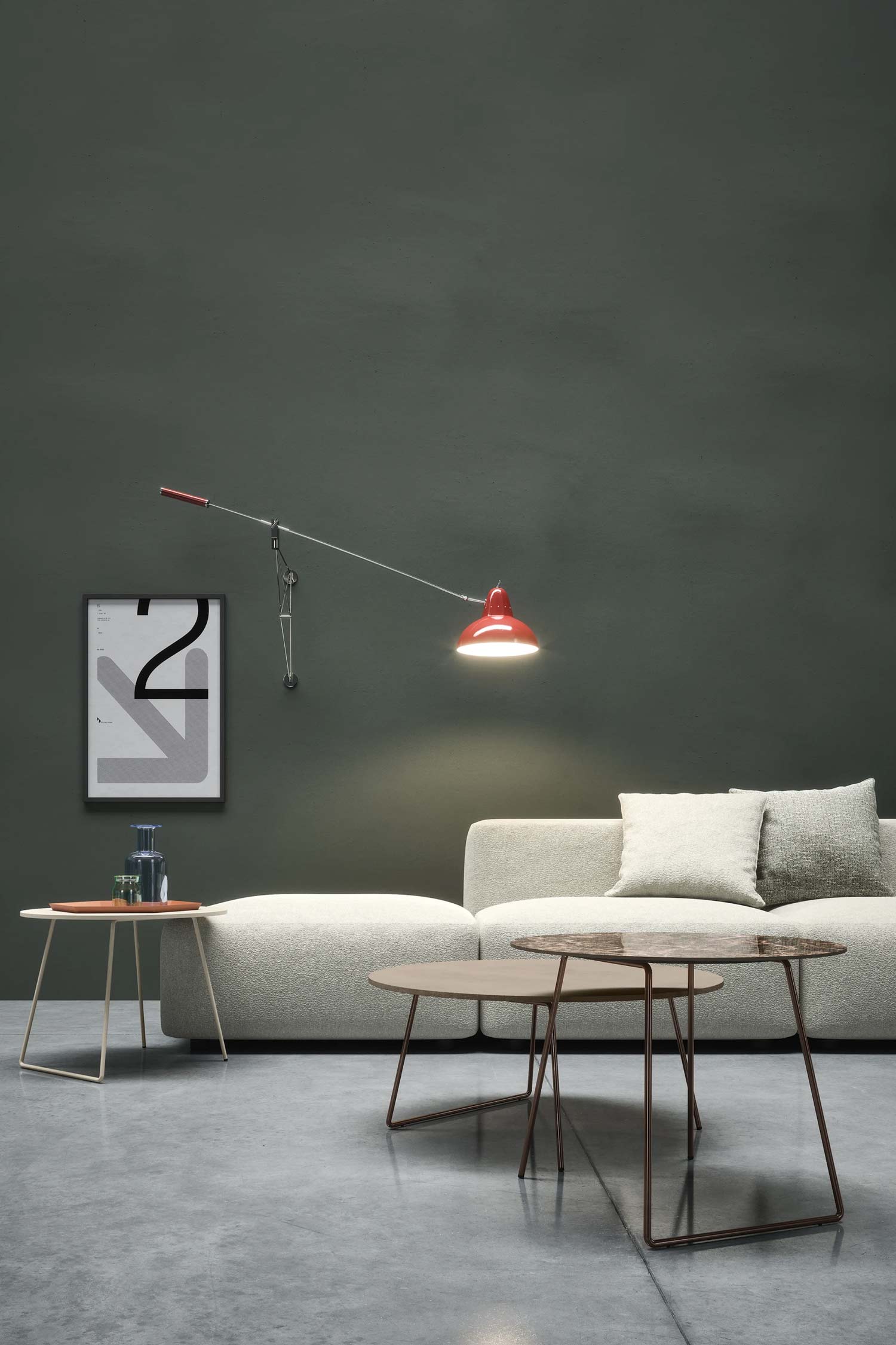 A vertical shot of a modern living room with a low-slung, cream-colored modular sofa against a dark gray-green wall. In the background, a swing-arm wall lamp with a red shade hangs over the couch. Two nesting coffee tables with bronze legs and light brown, rounded tops sit in front of the sofa. A smaller, light-colored side table with a red tray and glass decanter is to the left. A framed, abstract black and white print is on the wall behind it. The floor is a polished light gray concrete.