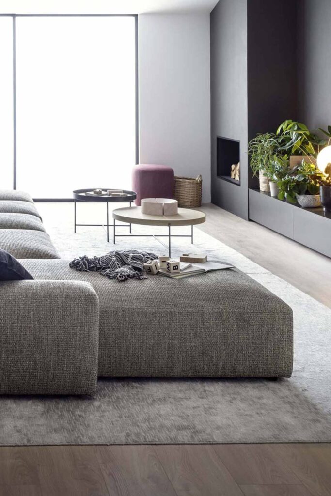 A vertical, close-up shot of a modern living room from the side of a large, gray modular couch. A dark gray and white throw blanket and a set of wooden letter blocks sit on a large ottoman section in the foreground. In the background, there is a built-in fireplace with a low media console and a collection of plants and lamps. A small pink ottoman, a round wooden coffee table, and a black coffee table are near the couch. A large window is on the left. The room features a gray rug over light-colored wooden floors.