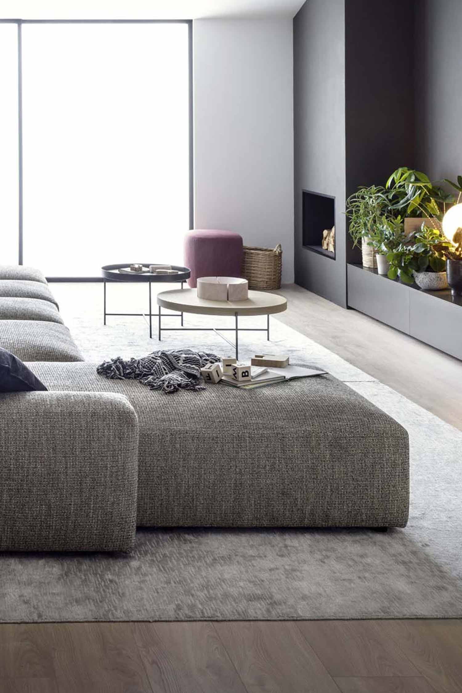A vertical, close-up shot of a modern living room from the side of a large, gray modular couch. A dark gray and white throw blanket and a set of wooden letter blocks sit on a large ottoman section in the foreground. In the background, there is a built-in fireplace with a low media console and a collection of plants and lamps. A small pink ottoman, a round wooden coffee table, and a black coffee table are near the couch. A large window is on the left. The room features a gray rug over light-colored wooden floors.