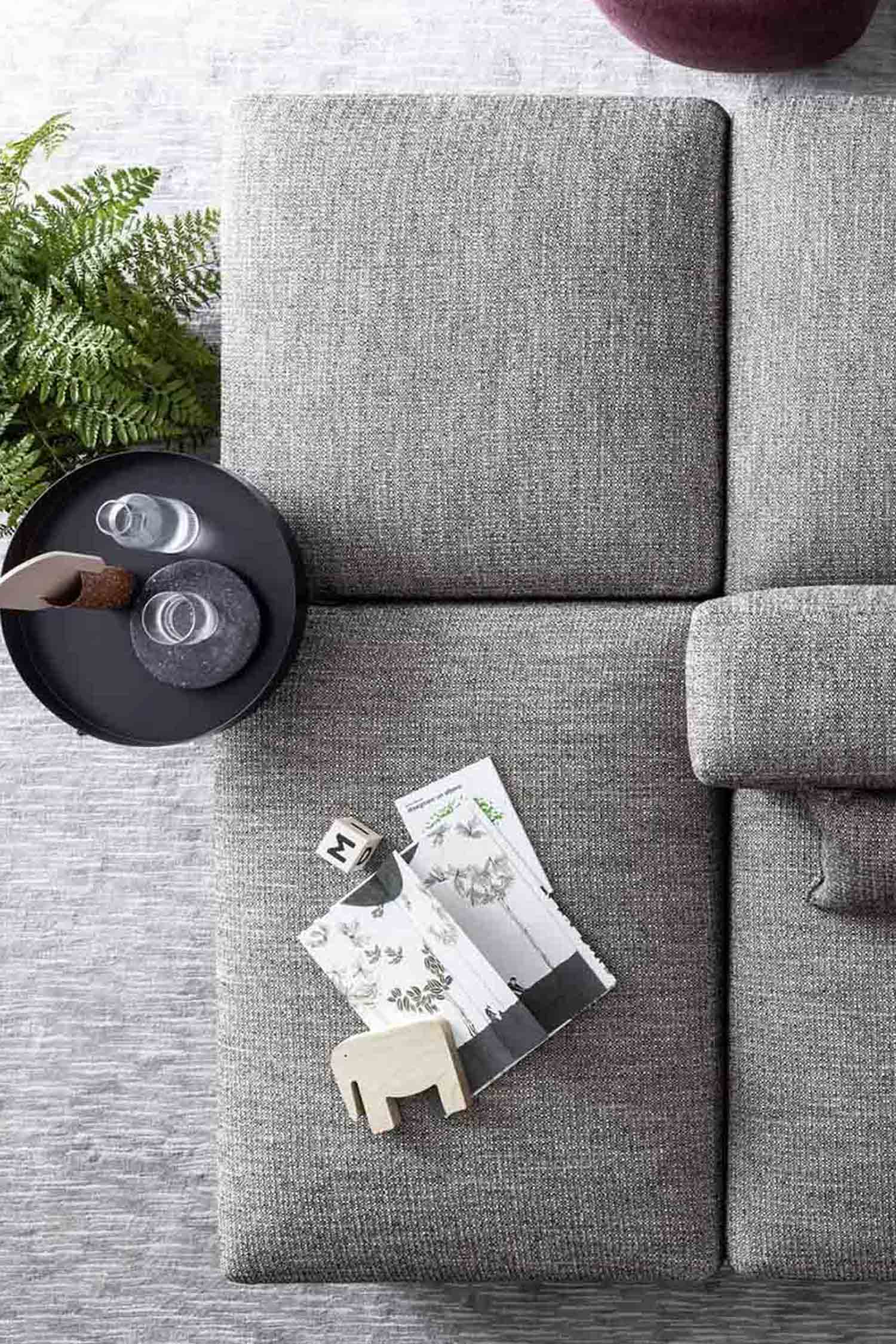 An overhead view of a light gray, textured modular couch. A black tray with two glasses and a coaster sits on the left seat, next to a small green plant. A small wooden block and a magazine with a white floral pattern are on a separate seat in the foreground.