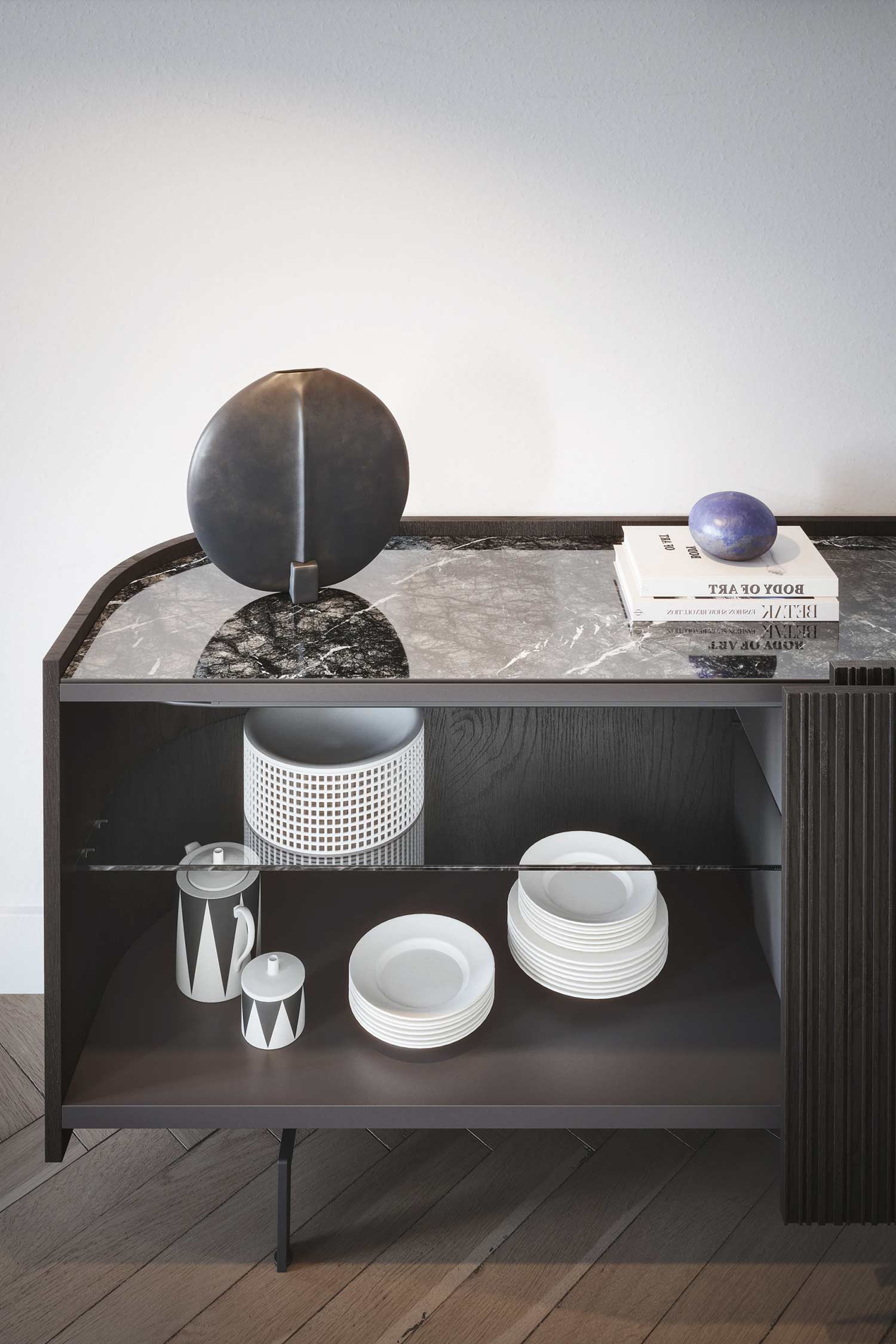 A close-up, three-quarter view of a dark wood and glass sideboard with a black marble top. The sideboard has a single, ribbed door on the right and an open section on the left with a clear glass shelf. On the top shelf, a black decorative object, a stack of white plates, and a patterned pitcher with a matching sugar pot are displayed. The top of the sideboard is adorned with a large, black sculpture, a decorative blue sphere, and a book. The floor is made of parquet wood.
