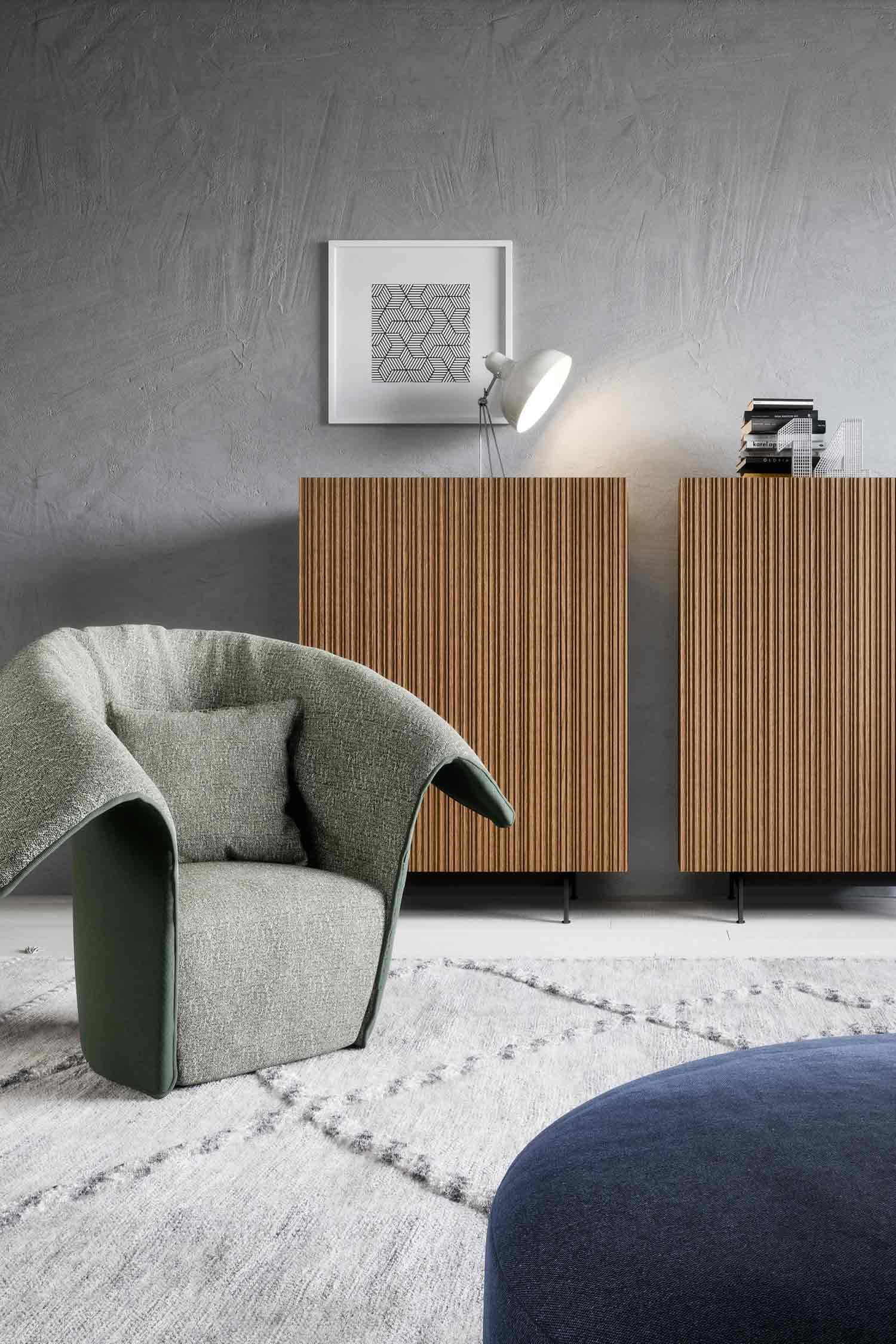 A stylish living room scene featuring two matching wood sideboards with a fluted texture, positioned side by side against a textured gray wall. Above the sideboards, a modern white desk lamp illuminates the area, and a framed geometric print hangs on the wall. In the foreground, a uniquely shaped, light gray and green upholstered armchair sits on a plush, patterned rug. A dark blue ottoman is partially visible on the right.