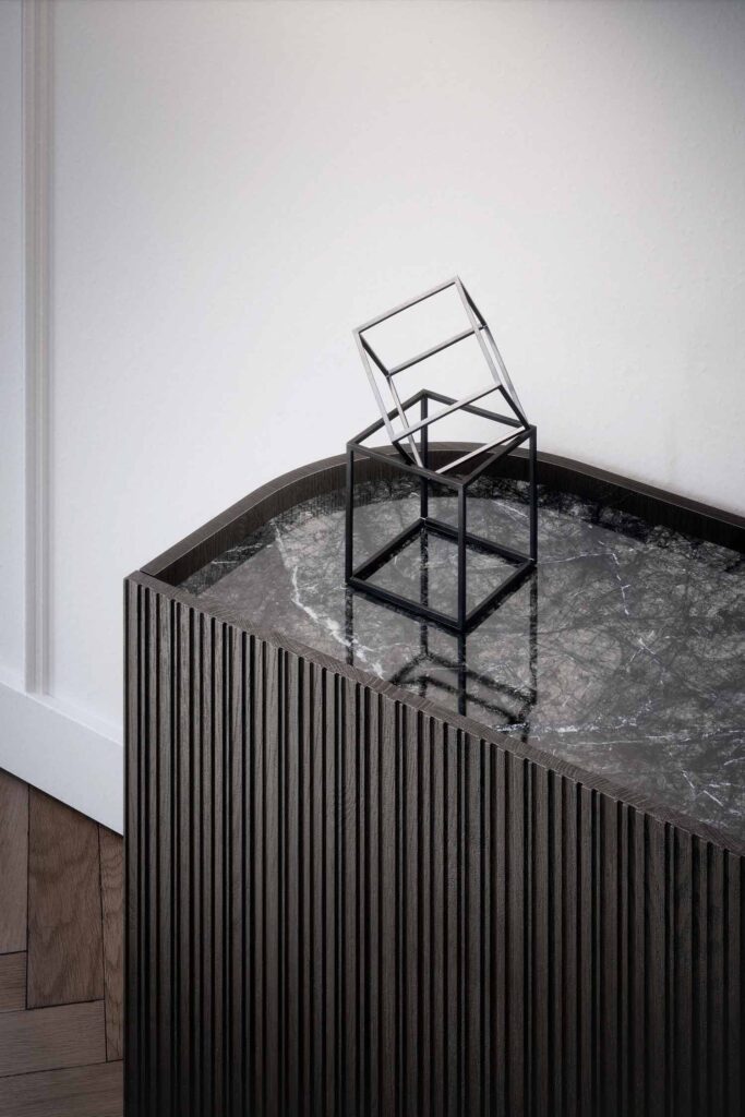A close-up, high-angle shot of the top of a modern dark-wood sideboard with a fluted texture. The sideboard has a dark gray marble top. A minimalist, sculptural black wire cube with another, smaller tilted cube inside sits as a decorative object on the marble surface. The background shows a simple white wall and a light wood parquet floor.
