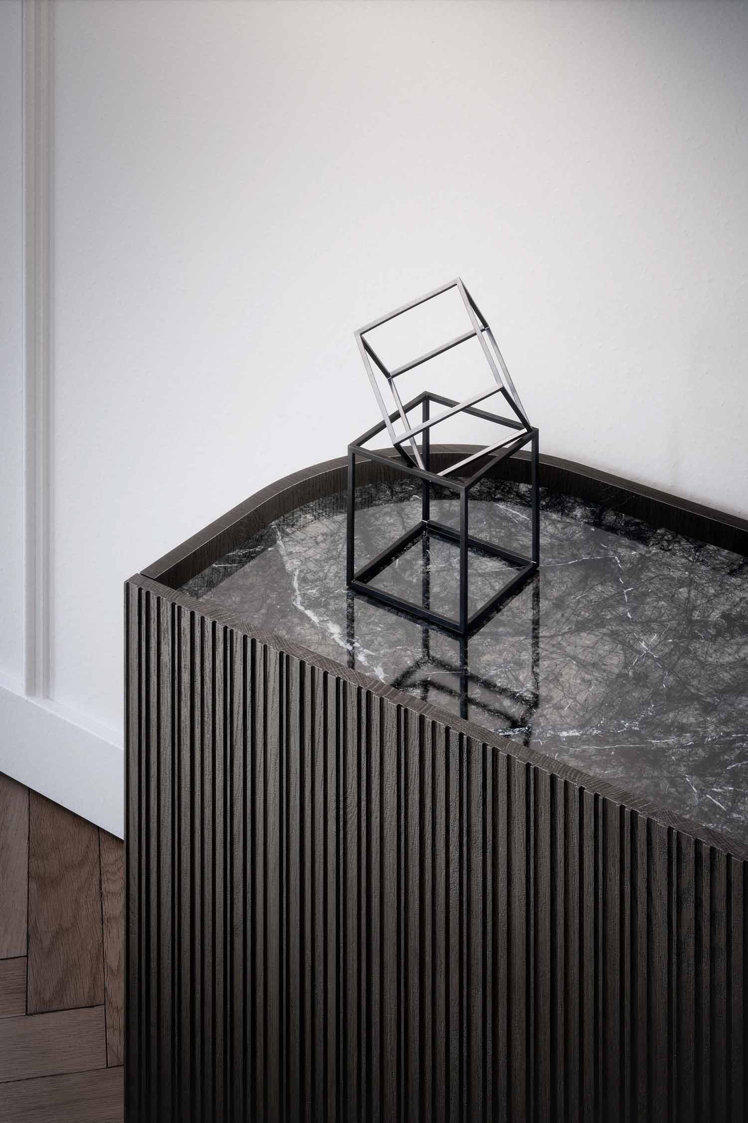 A close-up, high-angle shot of the top of a modern dark-wood sideboard with a fluted texture. The sideboard has a dark gray marble top. A minimalist, sculptural black wire cube with another, smaller tilted cube inside sits as a decorative object on the marble surface. The background shows a simple white wall and a light wood parquet floor.