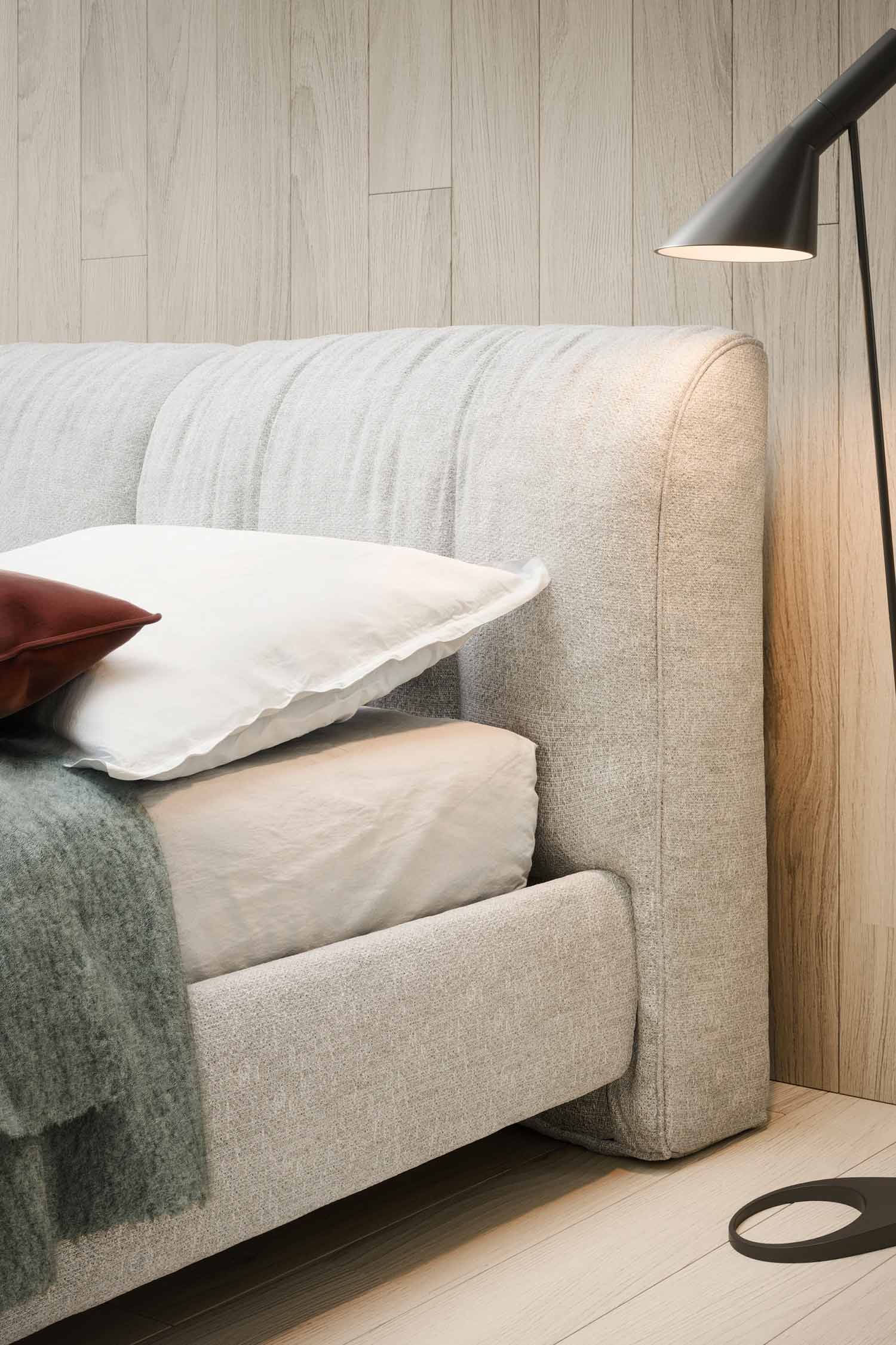 A close-up side view of the light grey upholstered bed frame and ribbed headboard. The textural details of the fabric are visible. A low, dark floor lamp stands next to the bed, which is placed against a vertical light wood-paneled wall.