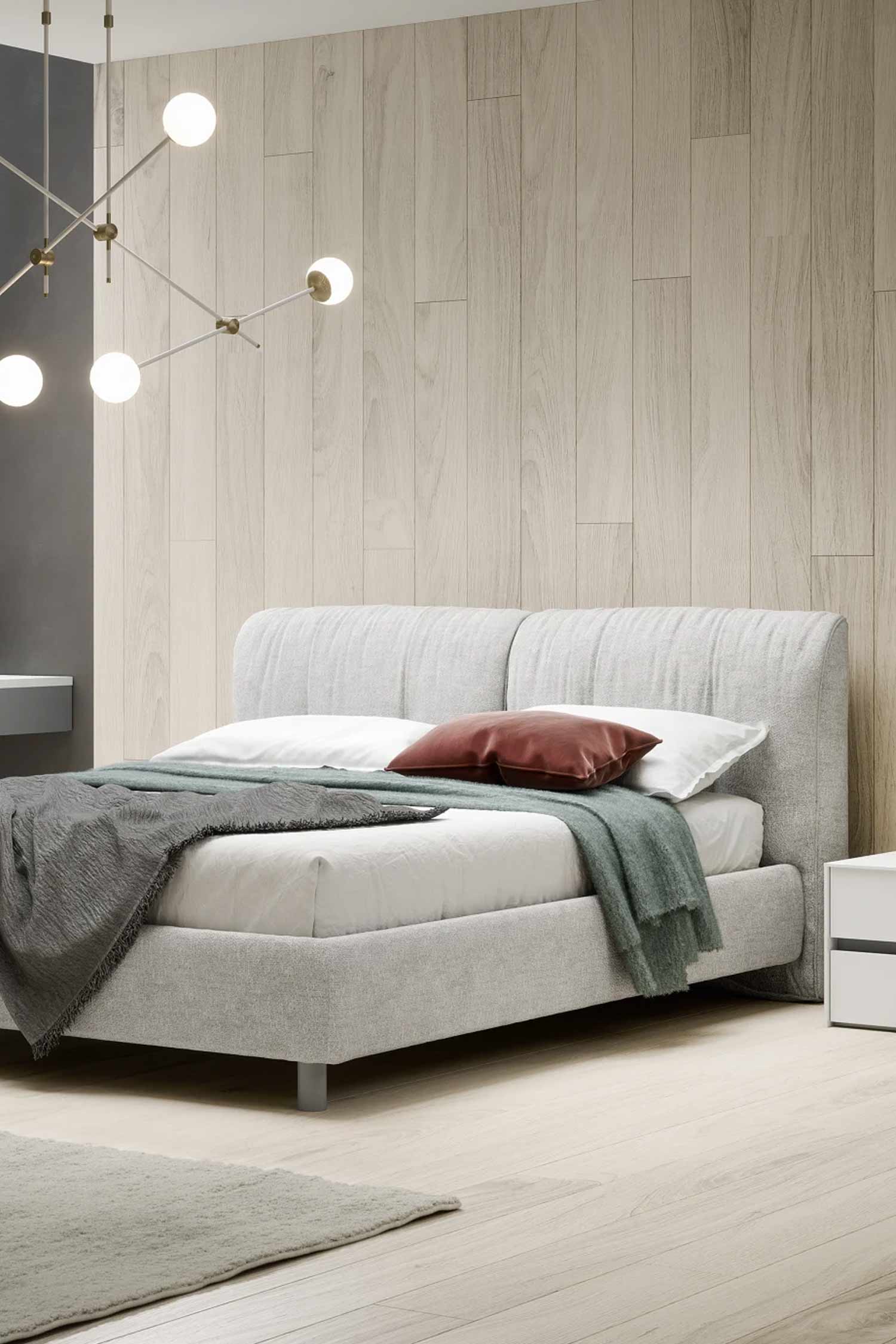 A light grey upholstered bed with a rounded, ribbed headboard and a low profile. The bed is dressed with a white sheet, a maroon pillow, and green and grey throw blankets. The bed is set against a light wood-paneled wall with a modern, spherical chandelier hanging above.