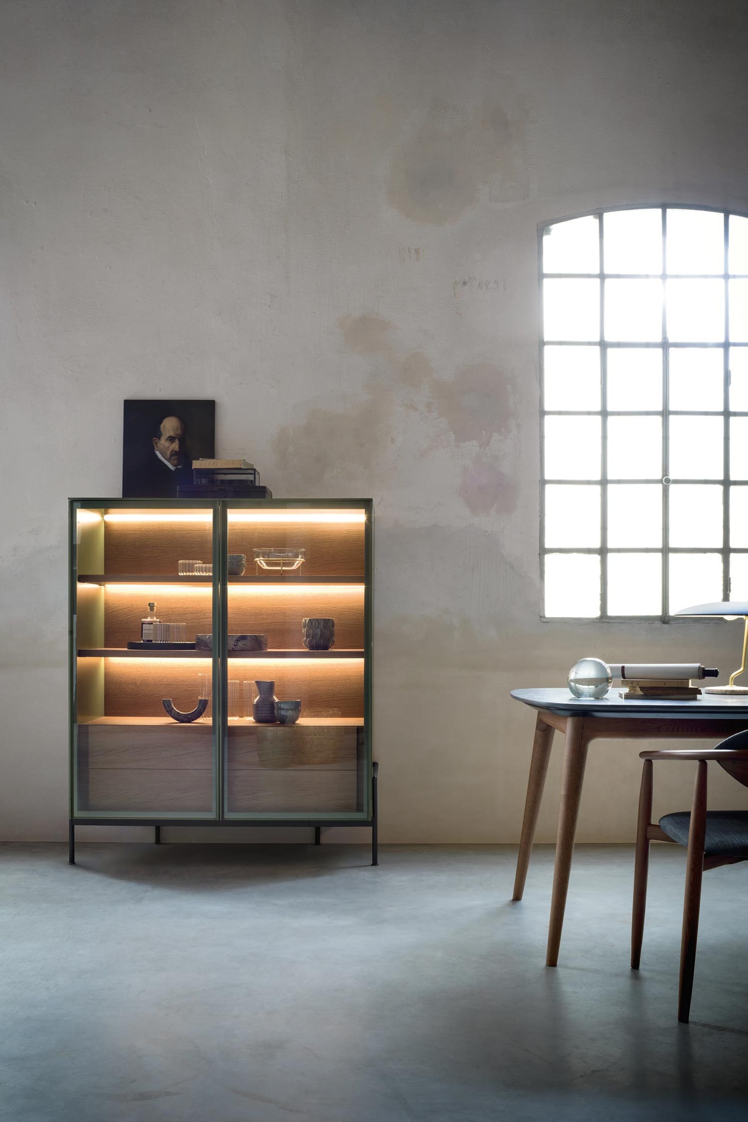 A modern, glass-fronted cabinet with a dark green frame and thin metal legs sits in a rustic room. The cabinet is illuminated from the inside with warm light, showcasing various decorative glass and ceramic objects on its shelves. To the right, part of a wooden table and a chair are visible, with a sunlit window in the background showing through its metal grid.