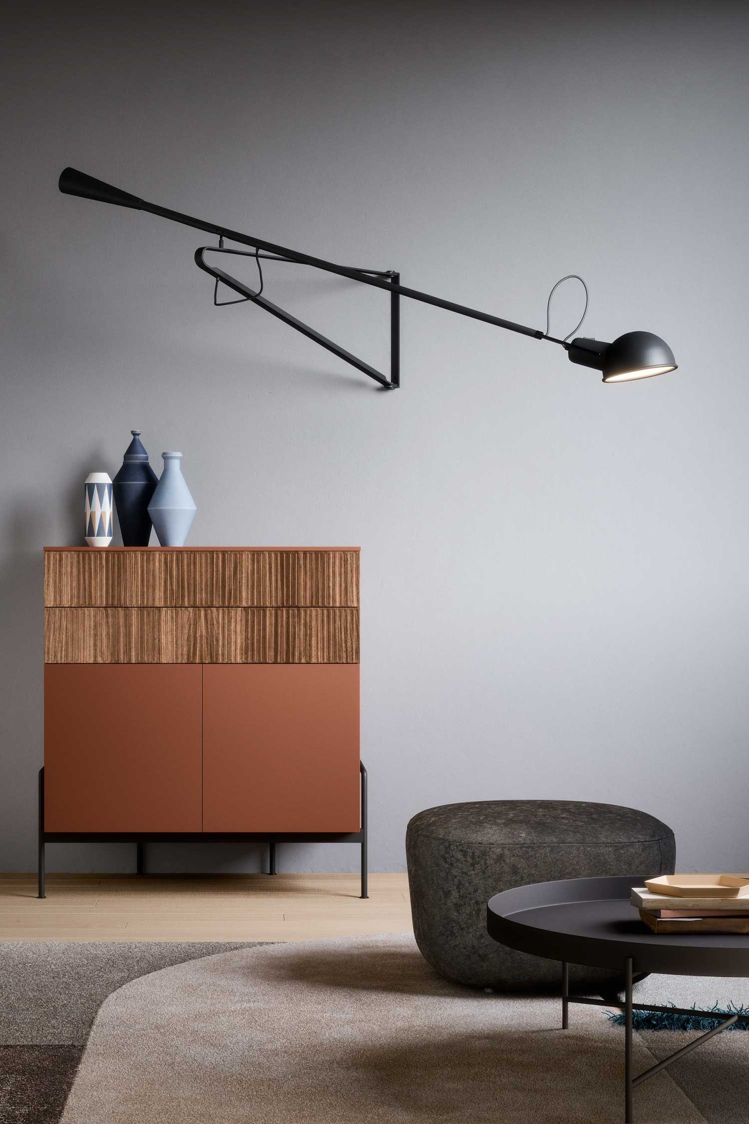 A high-angle, close-up shot of a modern room with a small, two-tone sideboard. The top half of the sideboard is made of natural wood with a textured, fluted pattern, while the bottom half has a smooth, reddish-brown finish. Three decorative vases in different shapes and colors sit on top. A long, black articulated lamp is mounted on the grey wall above the sideboard, and a textured, dark gray pouf is visible to the right.