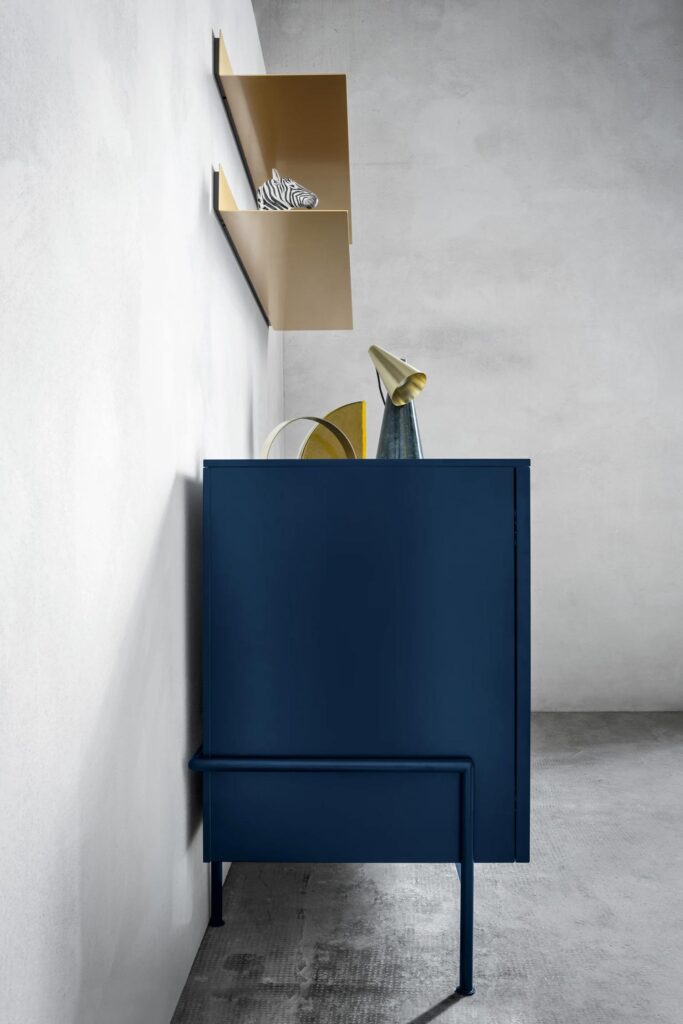 A side view of a minimalist, dark blue sideboard with a unique, open-bottom shelf design. A thin metal leg extends from the top of the sideboard down to the floor. The top of the sideboard holds a golden metal light fixture and a yellow decorative object. Above it, two gold-colored metal shelves are mounted on a rustic-looking concrete wall. A small zebra figurine is on the top shelf.