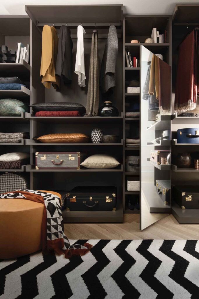A close-up, angled view of a luxurious walk-in closet. The dark gray shelving units are filled with a mix of hanging clothes, folded items, and decorative trunks. In the foreground, a large round ottoman is visible on a black and white chevron rug. A section of the closet with a reflective door is open on the right, revealing more shelving and a glimpse of the interior.