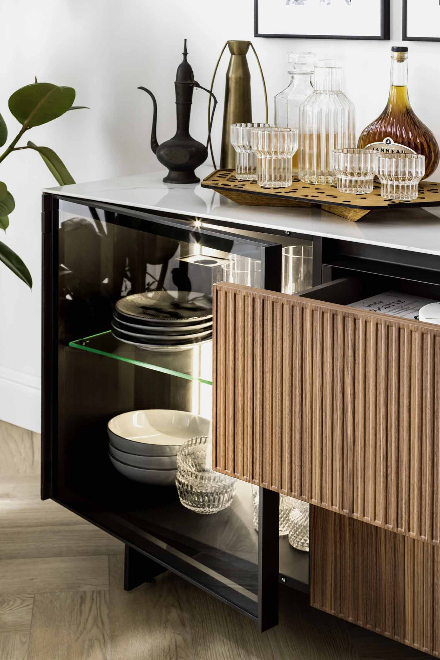 A close-up of a modern sideboard with a white marble top, featuring a dark wood frame. The left side has a clear glass door, revealing stacked plates and bowls on lit glass shelves. The right side has a light wooden fluted panel drawer that is slightly open, showing its contents. On top of the sideboard, a decorative black metal teapot, a gold vase, and a wooden tray with various glass bottles and drinking glasses are arranged. A green plant is partially visible on the far left.