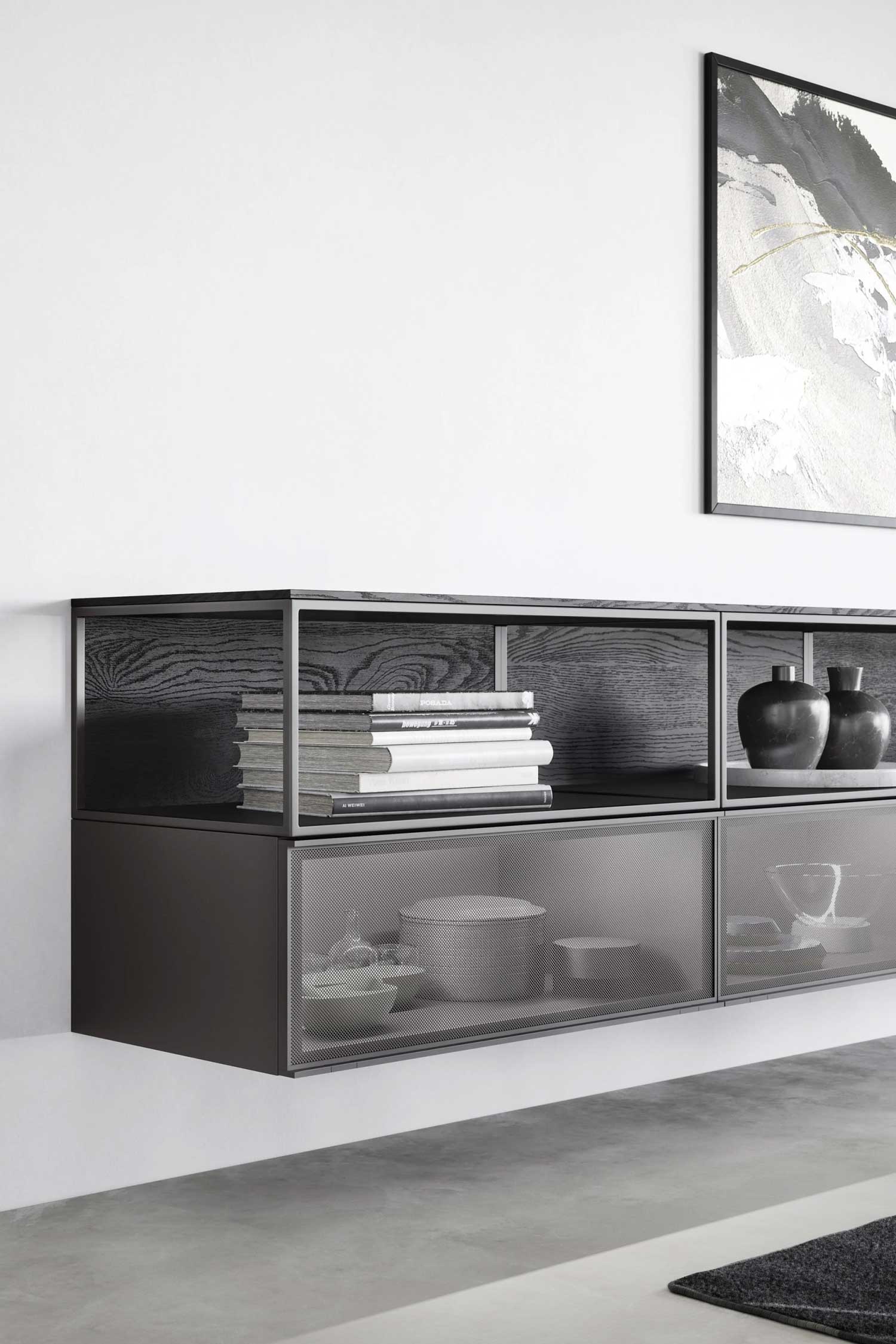 A close-up of a modern, floating sideboard with a dark frame and white interior, mounted on a white wall. The top section is open, with a dark wood grain back, and contains a stack of books and two black vases. The bottom sections are enclosed with a fine gray wire mesh, and through the mesh, various white and gray bowls are visible on the left, and a single decorative object on the right. A framed abstract painting with black and gold brushstrokes is partially visible on the wall in the upper right corner