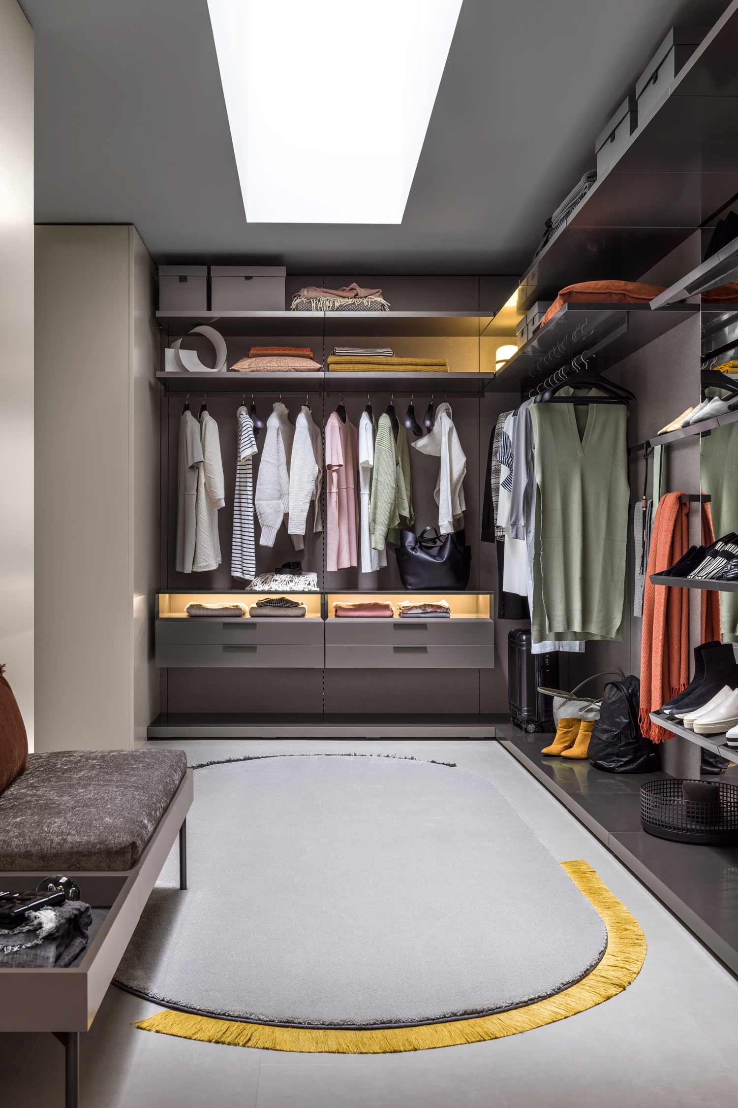 A wide-angle view of a modern walk-in closet with ambient lighting. Clothes are organized on a central hanging rack and on shelves above and to the sides. The floor has a gray oval rug with a bright yellow fringe. A bench with a textured cushion is in the foreground, and a suitcase and backpack are visible on the floor near a shoe shelf.