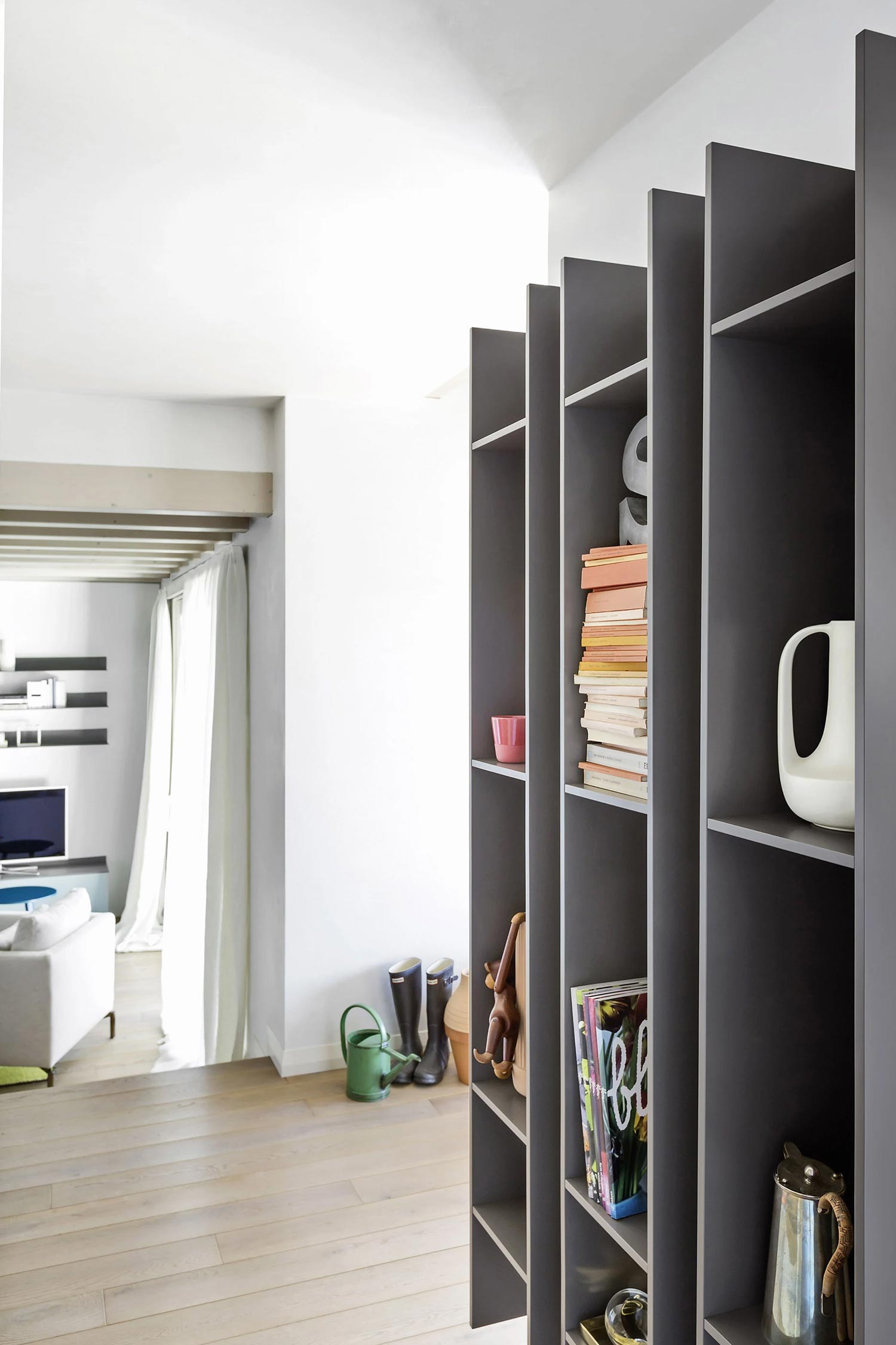 A close-up of a dark gray Butterfly luxury bookcase from Krieder studio, extending vertically from the floor to the ceiling. It serves as a room divider and holds books and decorative objects. The background shows a living area with a white sofa and a television.