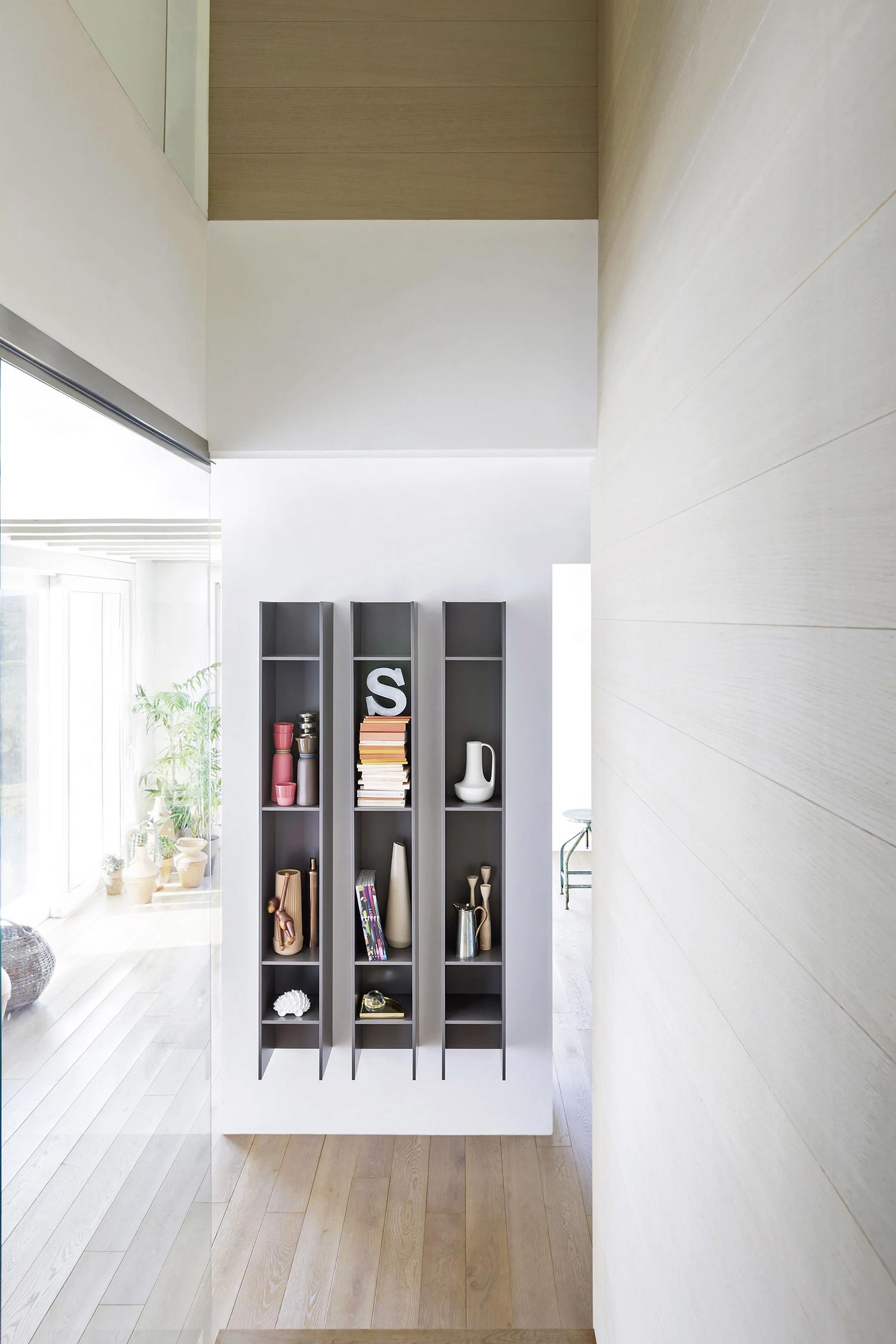 A shot of a wall with three tall, narrow Butterfly luxury bookcases from Krieder studio mounted on it. The dark gray shelving units are filled with a mix of books and various household decor, including a white pitcher and a large letter 'S'.