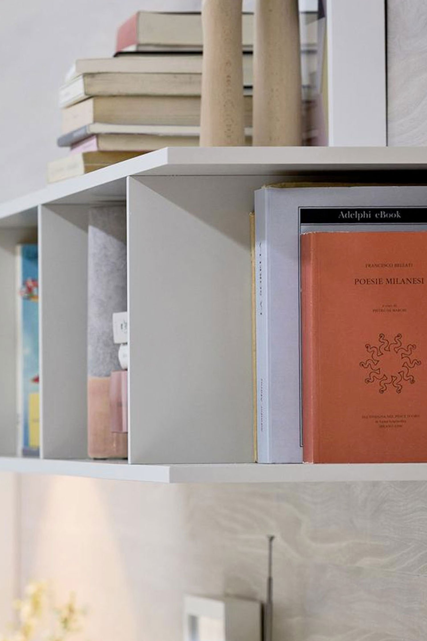A close-up of a white Butterfly luxury bookcase from Krieder studio, mounted on a wall. The shelf is filled with books, including an orange-covered one titled "Poesie Milanesi," and other items.