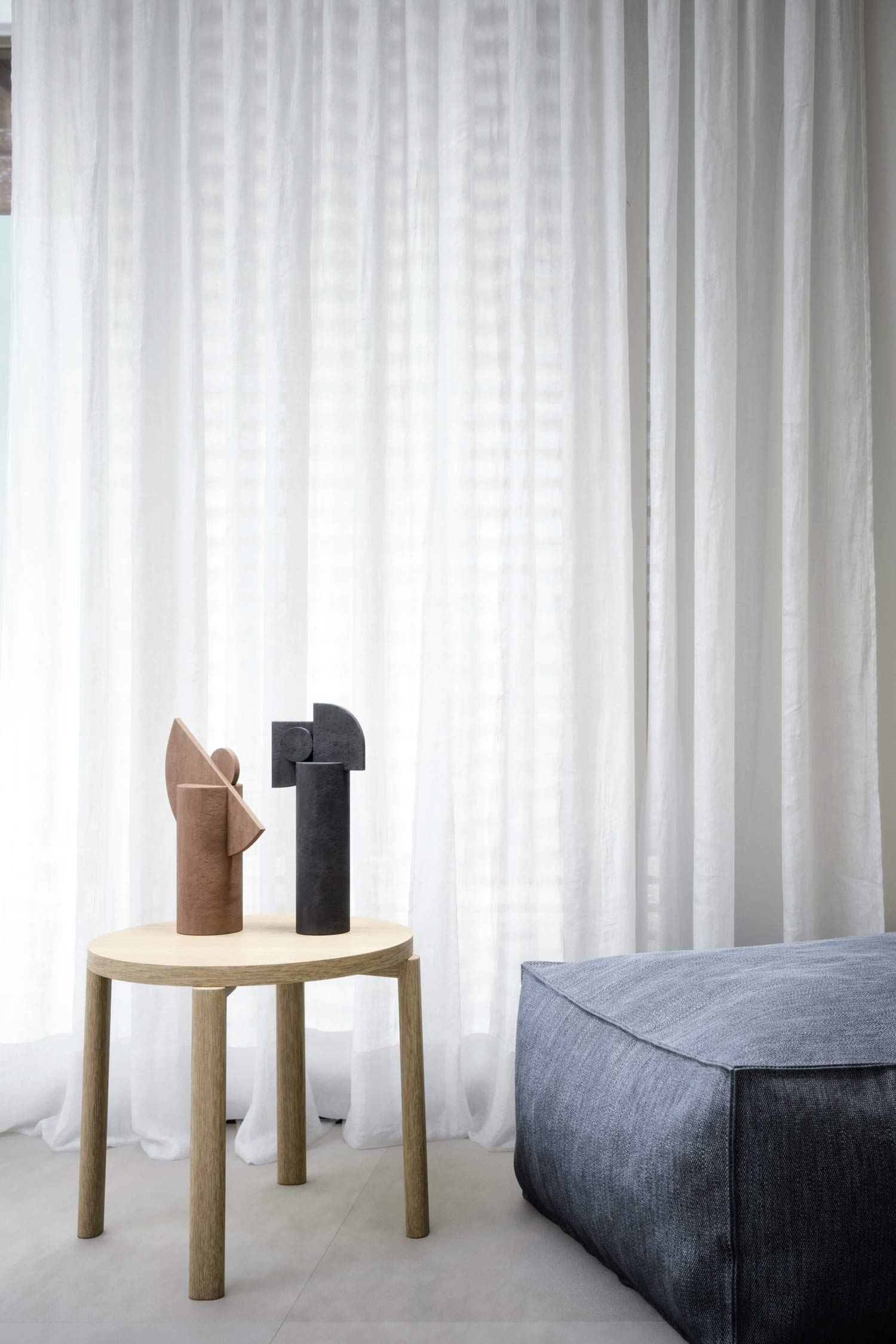 A modern interior features sheer white curtains in the background. In the foreground, a round light wood side table holds two abstract sculptural objects: one is terracotta-colored and the other dark gray. To the right, part of a large, rectangular dark gray fabric pouf is visible.