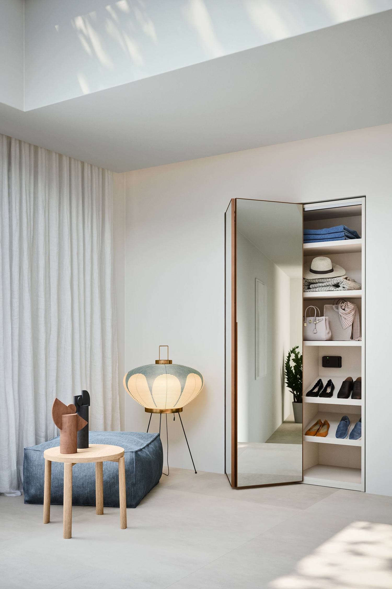 A stylishly decorated room with a minimalist aesthetic. On the left, a light wood side table with two abstract sculptural vases sits next to a large blue fabric pouf. A unique tripod floor lamp with a pleated lantern-style shade is placed behind the pouf. To the right, a large, tall, mirrored closet door is ajar, revealing an open shelving unit with shoes, clothes, and accessories neatly organized.
