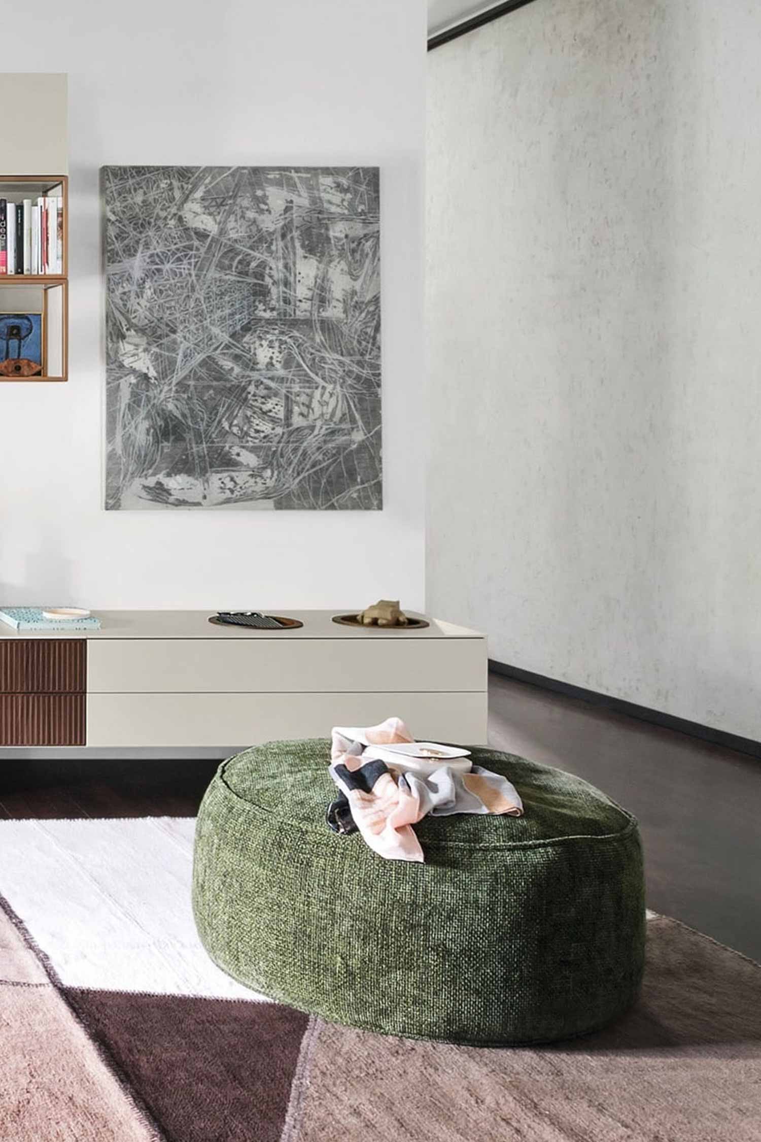 A large, textured round green fabric ottoman is in the foreground, with a folded scarf resting on top. The ottoman sits on a geometric patterned rug with colors including white, beige, and maroon. Behind the ottoman is a low, light-colored cabinet with two drawers, one with a wood-grain texture and the other with a dark ribbed texture. A large abstract gray and white painting hangs on the wall above the cabinet.
