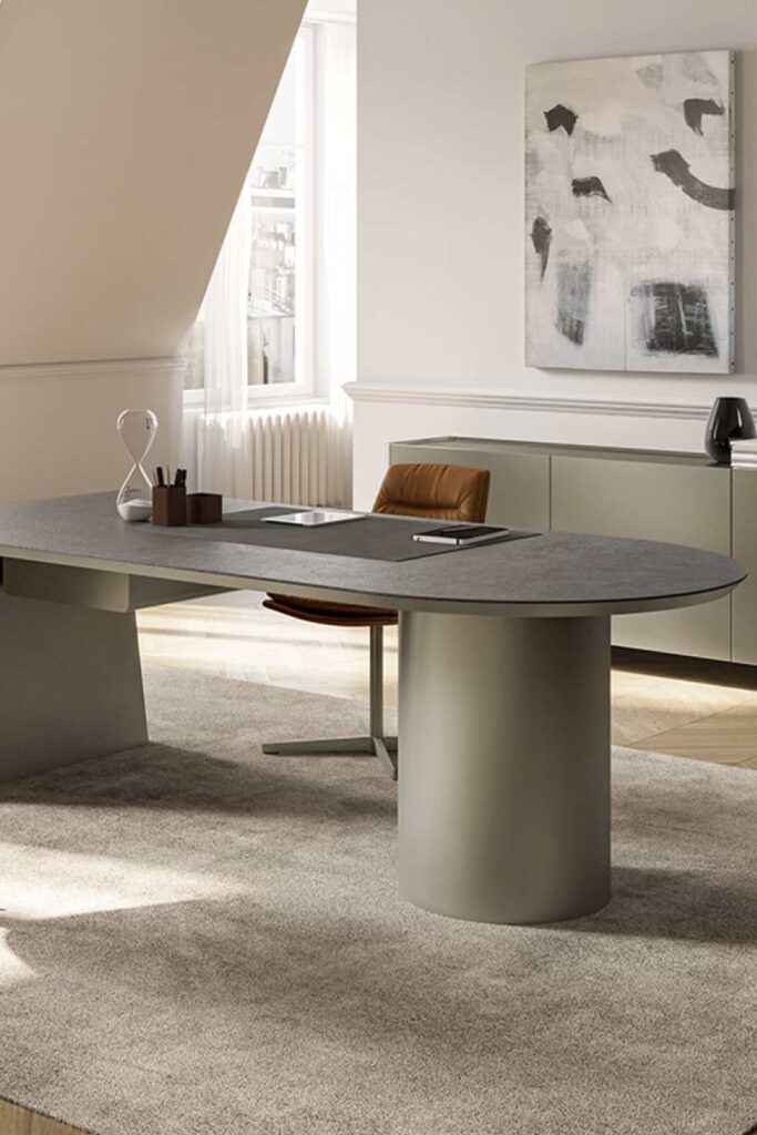A close-up shot of the matte gray Cilindro luxury desk in an office setting. The desk features an elliptical top resting on a thick, cylindrical base and a tapered pedestal leg. A brown leather chair is tucked under the desk, and an hourglass and desk mat are visible on the surface.