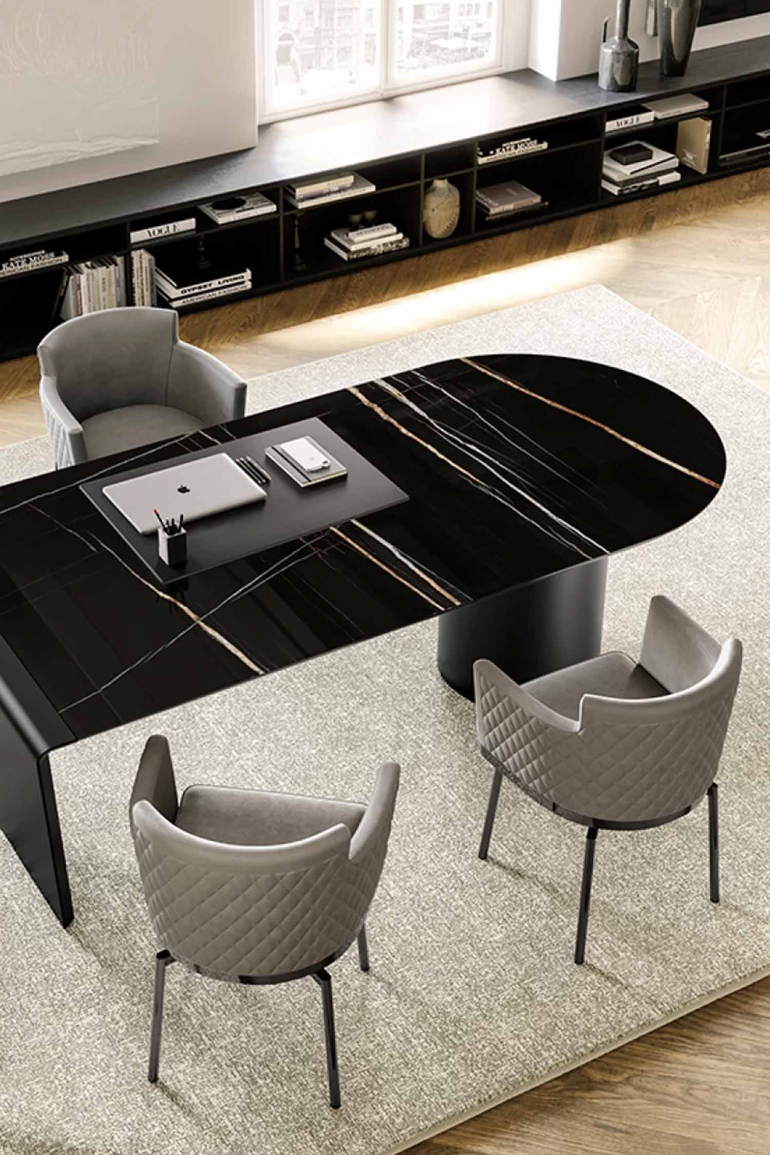 High-angle shot of the long, black marble-look Cilindro desk showing its sleek, rounded design and gold/white veining. Three gray upholstered chairs surround the desk, which sits on a large, textured rug. A laptop and desk pad are centered on the tabletop.
