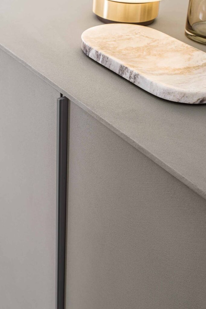 A close-up of the top and front of a modern, two-door cabinet with a textured, matte gray surface. A light-colored marble serving board rests on the top surface, along with the corner of a gold-toned decorative object. The cabinet features a thin, vertical black handle on the left door.