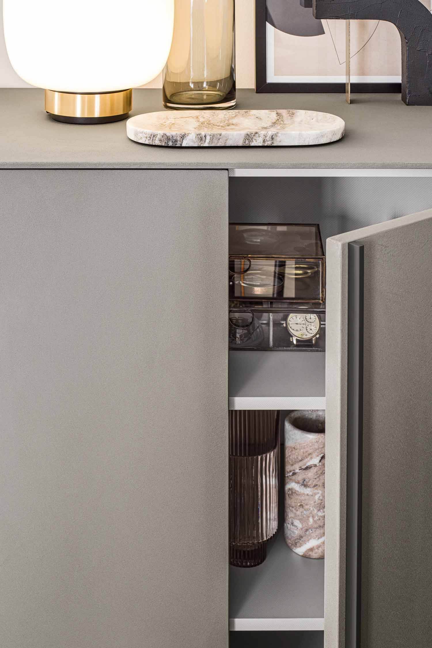 A close-up shot of a modern gray cabinet with a door slightly ajar, revealing the interior shelves. The cabinet's surface has a slight texture. Inside, the top shelf holds a clear acrylic box containing watches and other small items. The bottom shelf holds two decorative items: a ribbed glass vase and a round marble vase with pink and white tones. On top of the cabinet, a white and brass lamp, a brown glass vase, a marble tray, and a framed piece of art are visible.