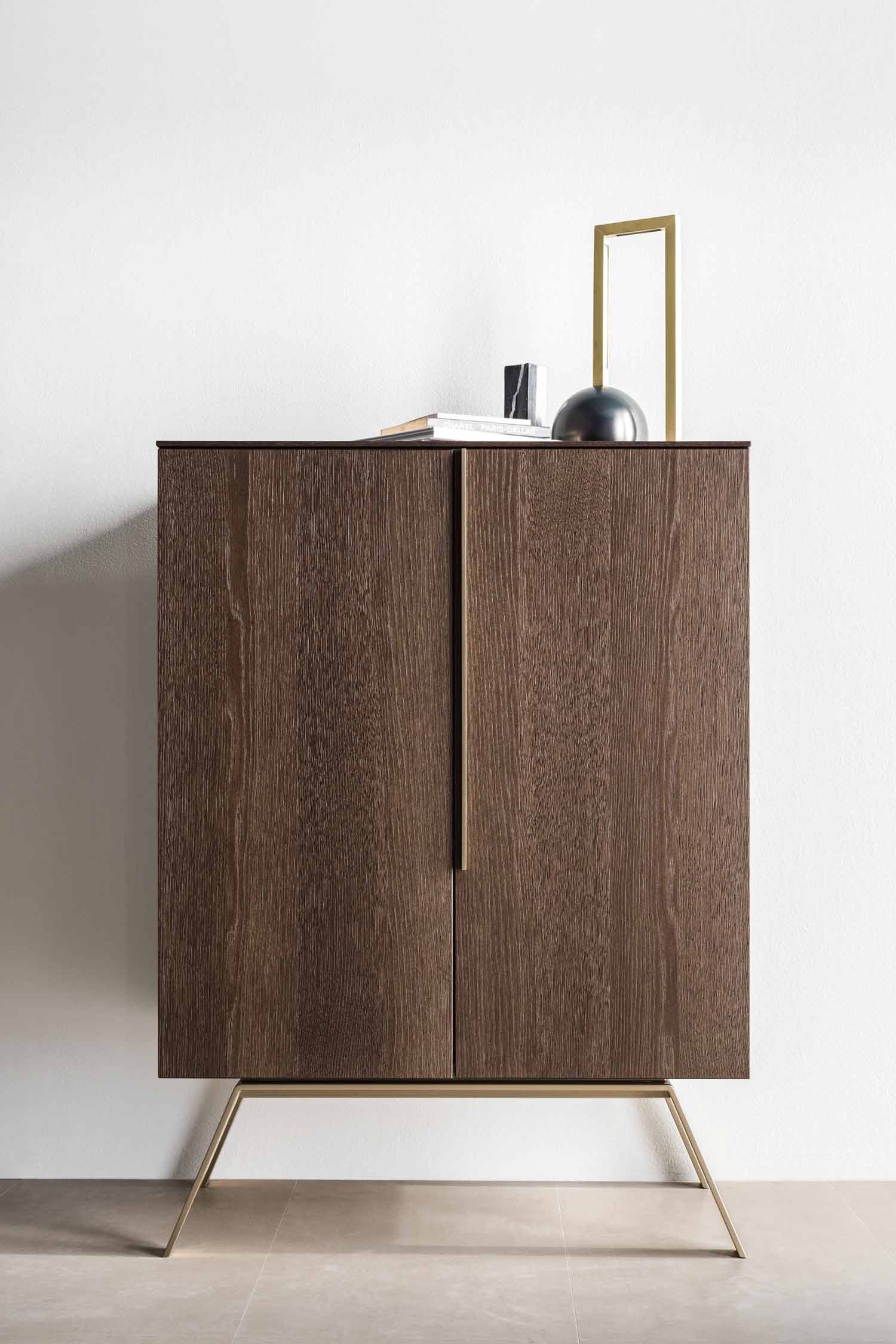 A stylish, modern two-door cabinet with a dark wood grain finish. The cabinet is supported by unique, slender, angled brass legs that give it a sophisticated look. A thin, black vertical handle is centered where the two doors meet. On the top surface, a few books and two decorative objects—a black dome-shaped item and a minimalist brass frame—are placed. The cabinet stands against a simple white wall.