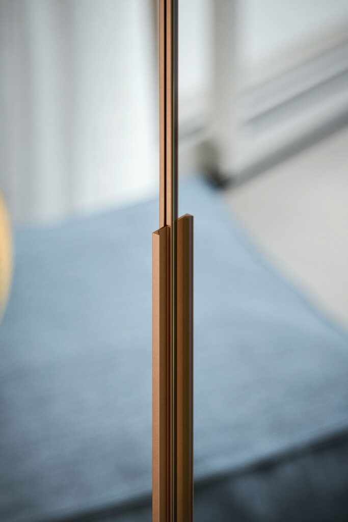A close-up vertical shot of a thin, metallic detail on a hinged wardrobe door. The object, which could be part of a handle or frame, is a polished rose gold or copper color. The background is a blurry light blue cushion.