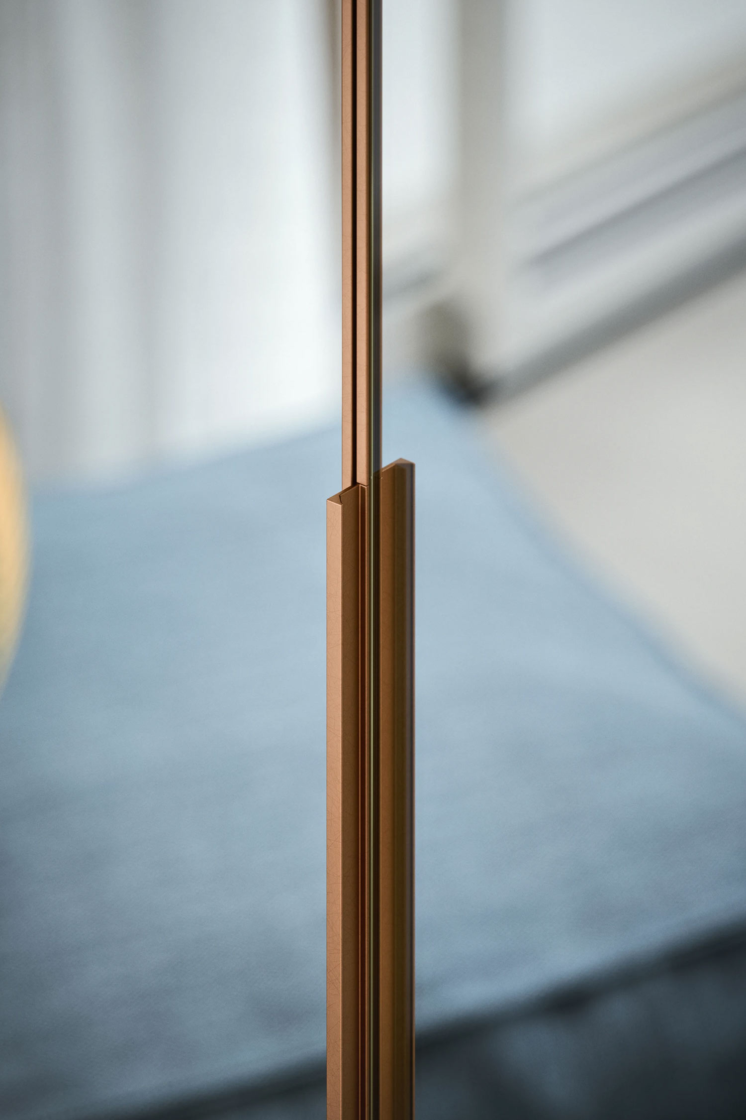 A close-up vertical shot of a thin, metallic detail on a hinged wardrobe door. The object, which could be part of a handle or frame, is a polished rose gold or copper color. The background is a blurry light blue cushion.