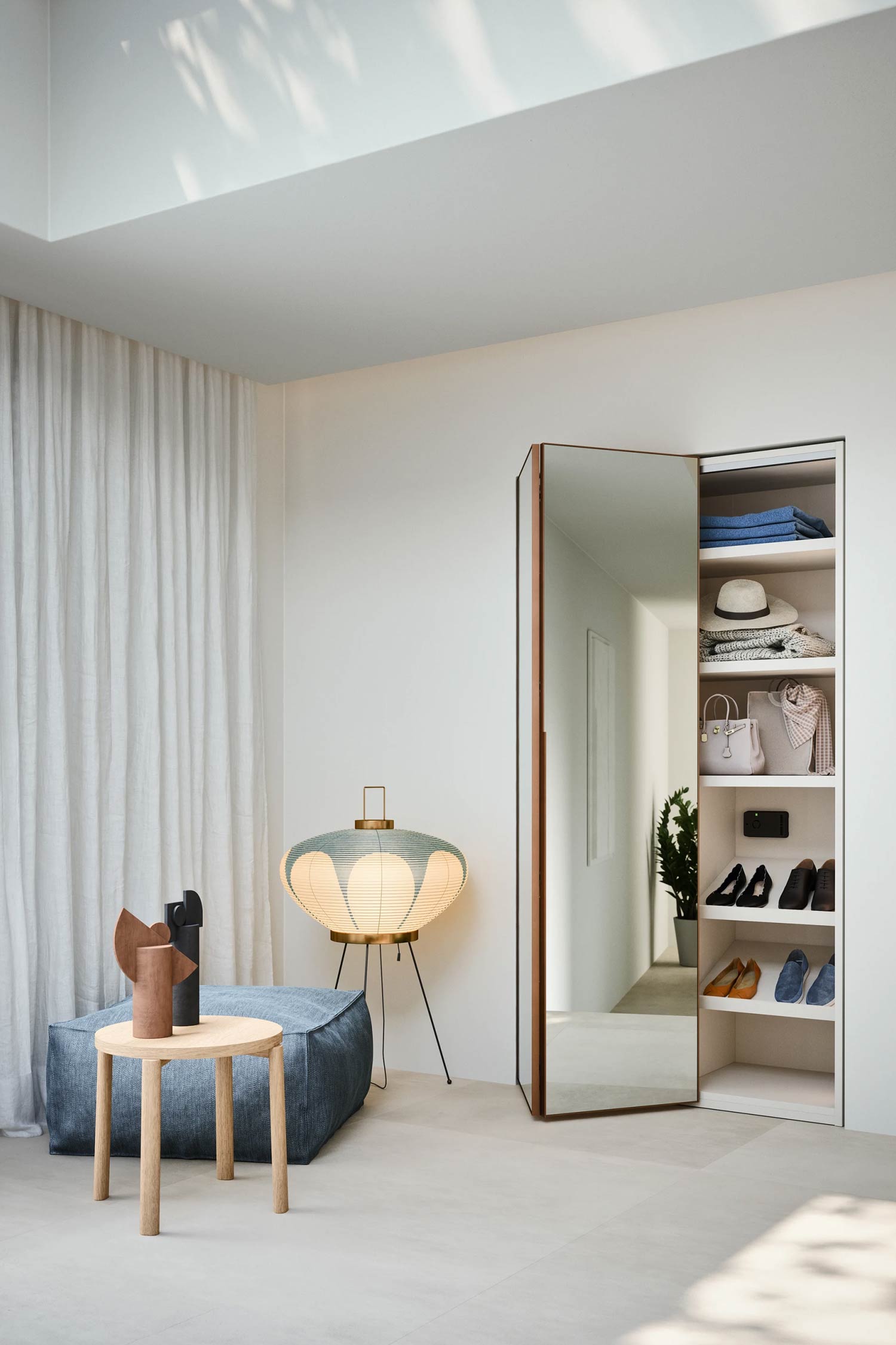A luxury hinged wardrobe with an open mirrored door that reveals a white shelved interior. The shelves hold various items like shoes, bags, a hat, and folded clothes. The room is bright and minimalist, featuring a blue pouf, a small wooden side table with decorative vases, and a large, unique, patterned lamp. The floor is a light-colored tile.