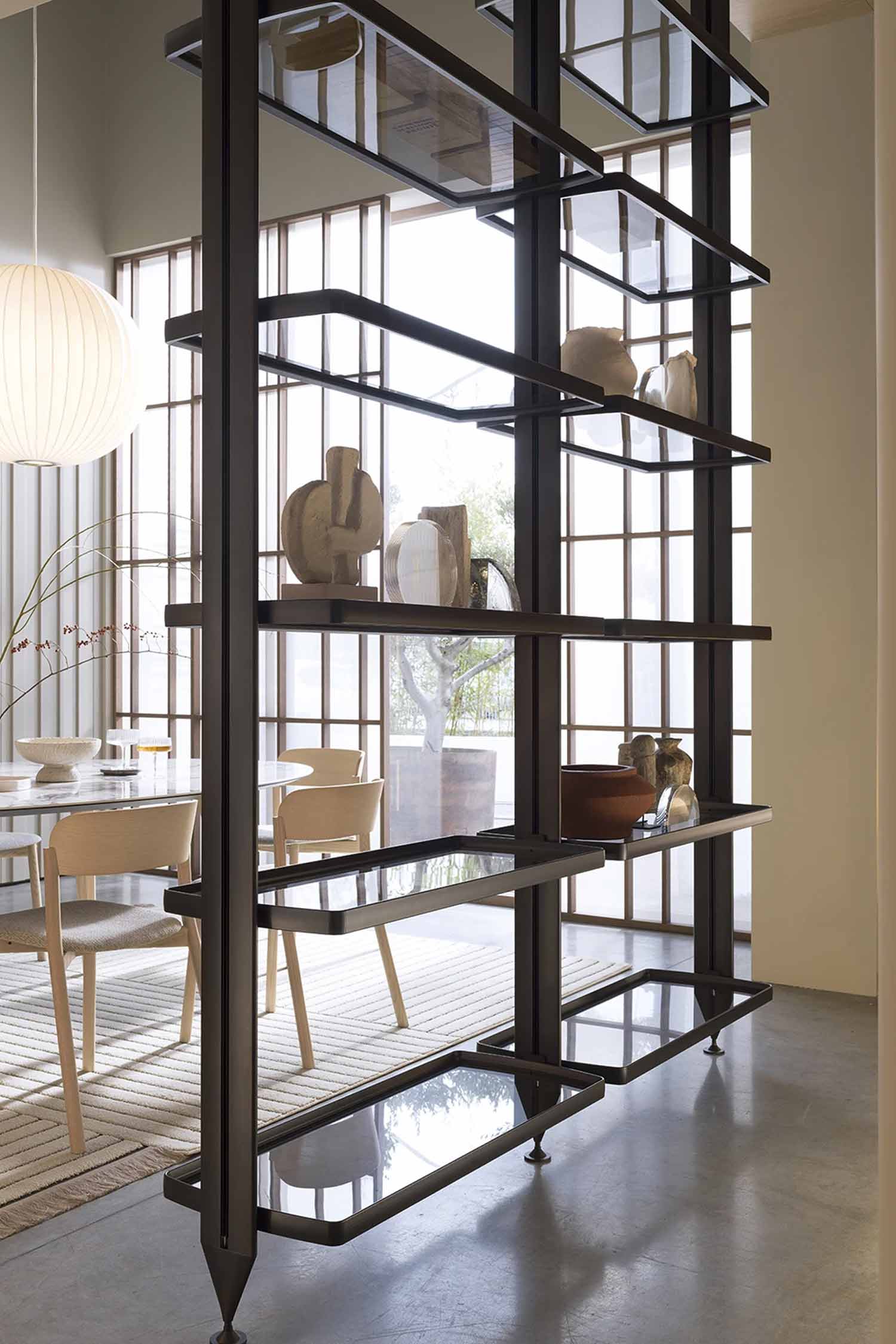 A modern, open shelving unit serves as a room divider. It is made of dark metal with clear glass shelves of varying lengths, creating an asymmetric, multi-level design. The shelves hold several pieces of abstract and minimalist pottery in neutral tones. In the background, a dining table with chairs and a large paper lantern hang in front of a window with a grid pattern.