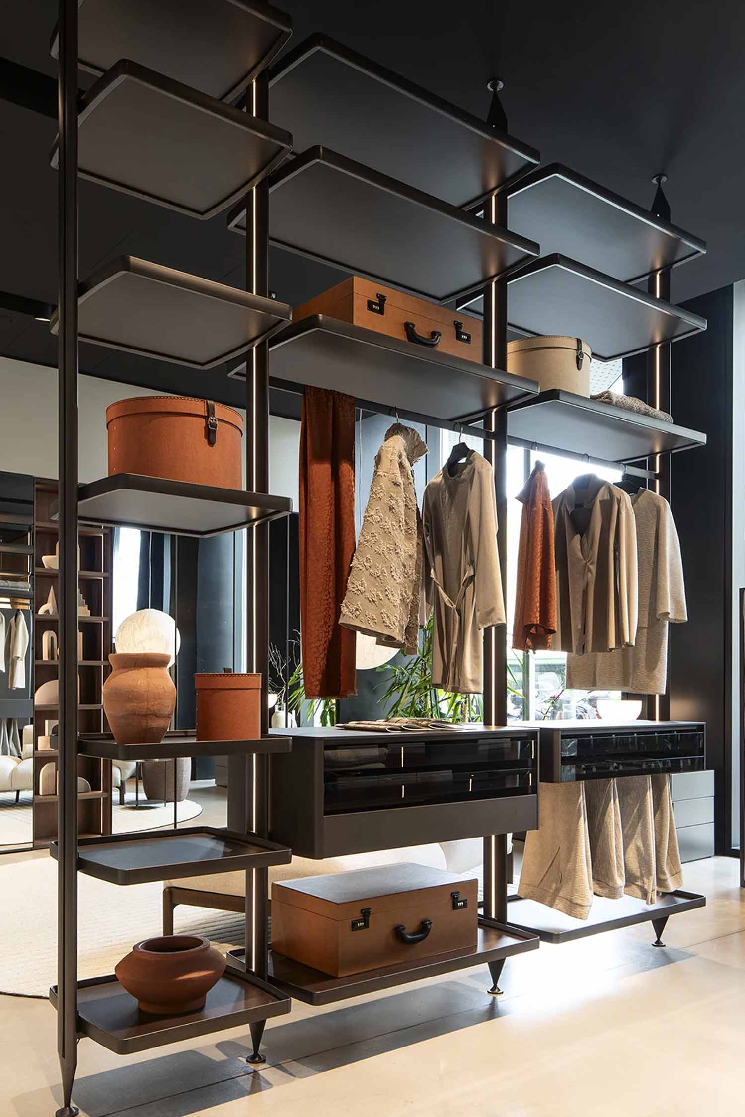 A modern walk-in closet or dressing room system is shown with a modular black metal frame and various shelves. The shelves hold a mix of clothing on hangers, terracotta-colored storage boxes and suitcases, and decorative vases. The system also includes a dark grey cabinet section with drawers. The overall style is sophisticated and minimalist, combining storage with display.