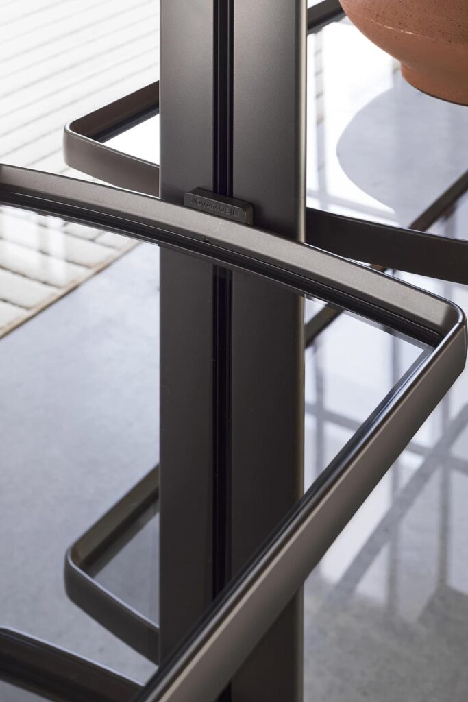 A close-up shot focuses on the detail of a modern modular shelving system. A vertical dark metal pole runs through the center, with a small "NOVAMOBILI" tag visible. Several horizontal dark metal frames, some holding clear glass shelves, extend from the pole. The sleek design and material textures are highlighted, with a blurred background suggesting other elements of the furniture.