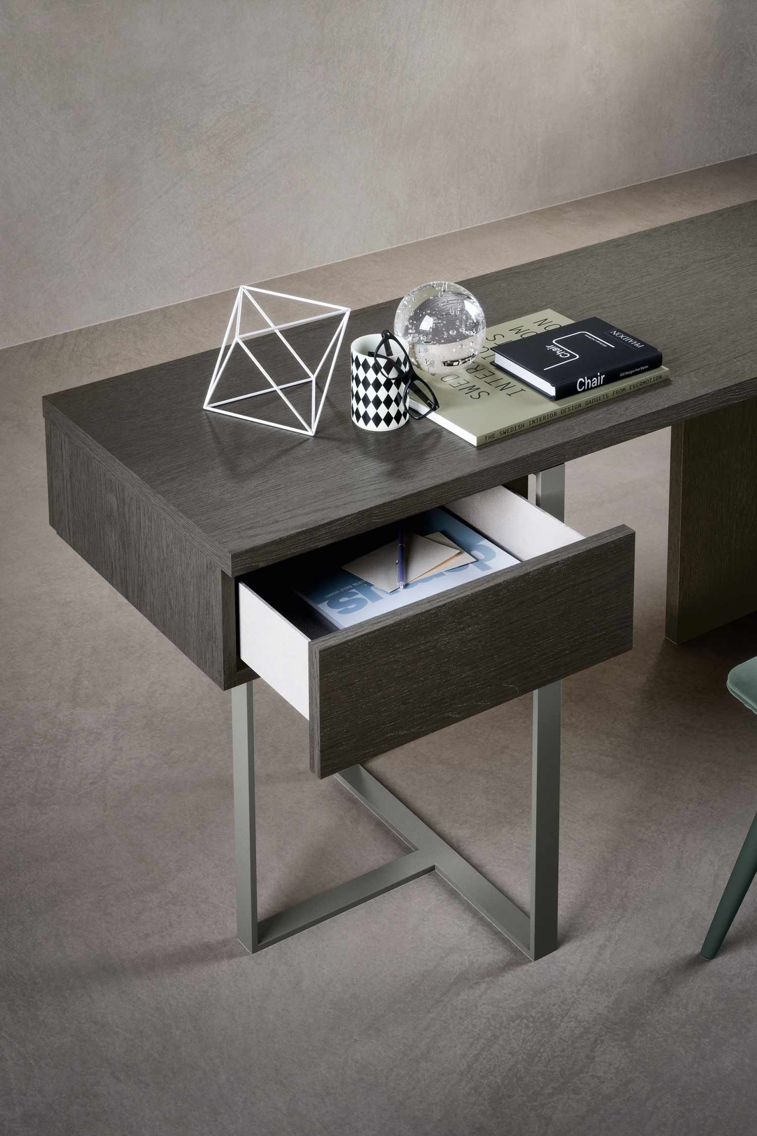 A detailed, high-angle shot of a dark wood and metal desk. The top of the desk holds a book titled Chair, a checkered mug, a pair of glasses, and a clear glass orb. A single, wide drawer is pulled out to reveal a white interior with a pen and various papers.