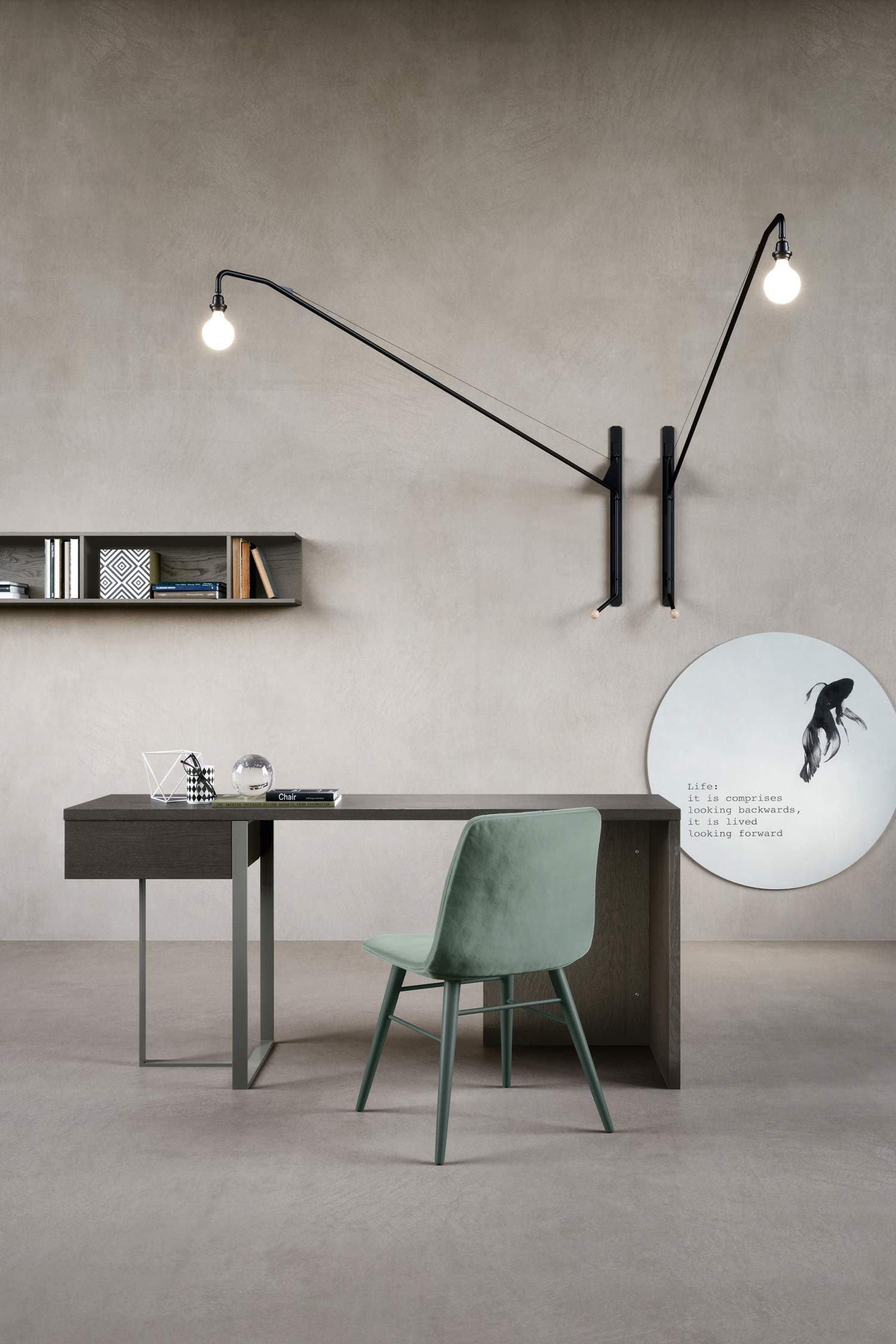 A minimalist office space featuring a dark wood desk with metal legs, a green velvet chair, and a round mirror with a fish motif and text. Above the desk is a long, black, adjustable wall lamp with two exposed bulbs. To the left, a floating shelf holds books and decorative items. The walls and floor are a neutral concrete or plaster finish.