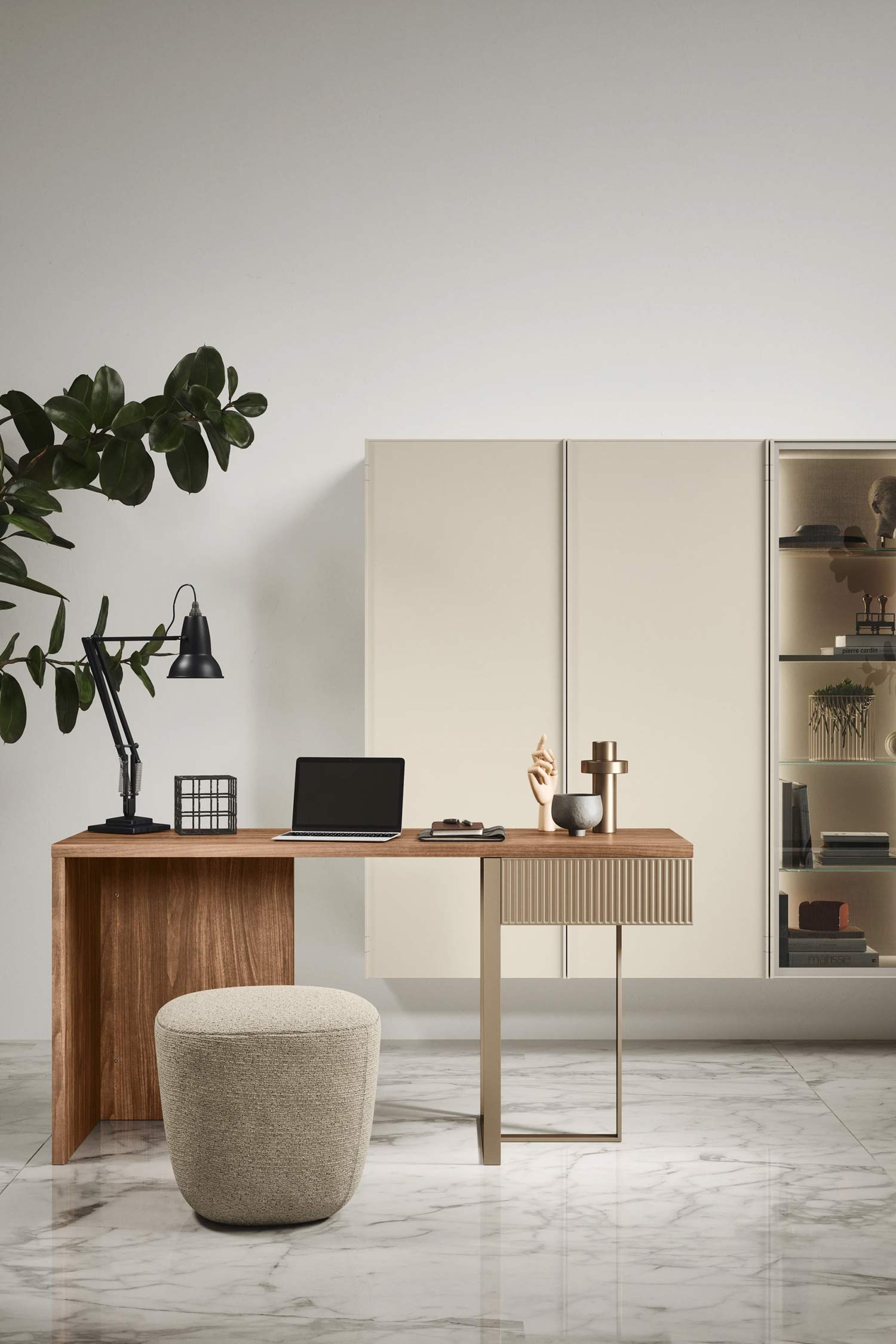 A bright, minimalist office space with a light wood desk and metal legs. On the desk are a laptop, a lamp, and some decorative objects. An upholstered, round stool is pulled up to the desk. In the background is a white cabinet with glass shelving on the right, and a large green plant on the left. The floor is white marble tile.