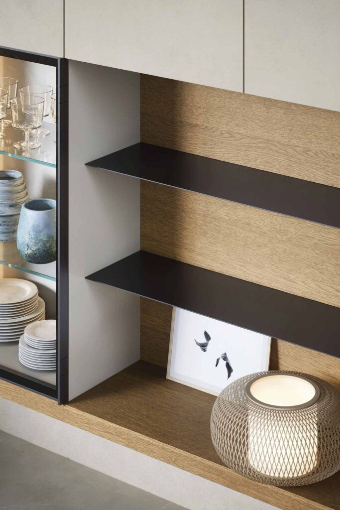 A close-up of a modern, built-in kitchen or dining room cabinet and shelving unit. The unit features a glass-front display case on the left with lighting, filled with various dishes, glassware, and a blue decorative vase. To the right, open shelving with a light wood back panel and dark gray metal shelves holds a framed abstract print and a decorative lamp with a textured, metallic mesh shade.