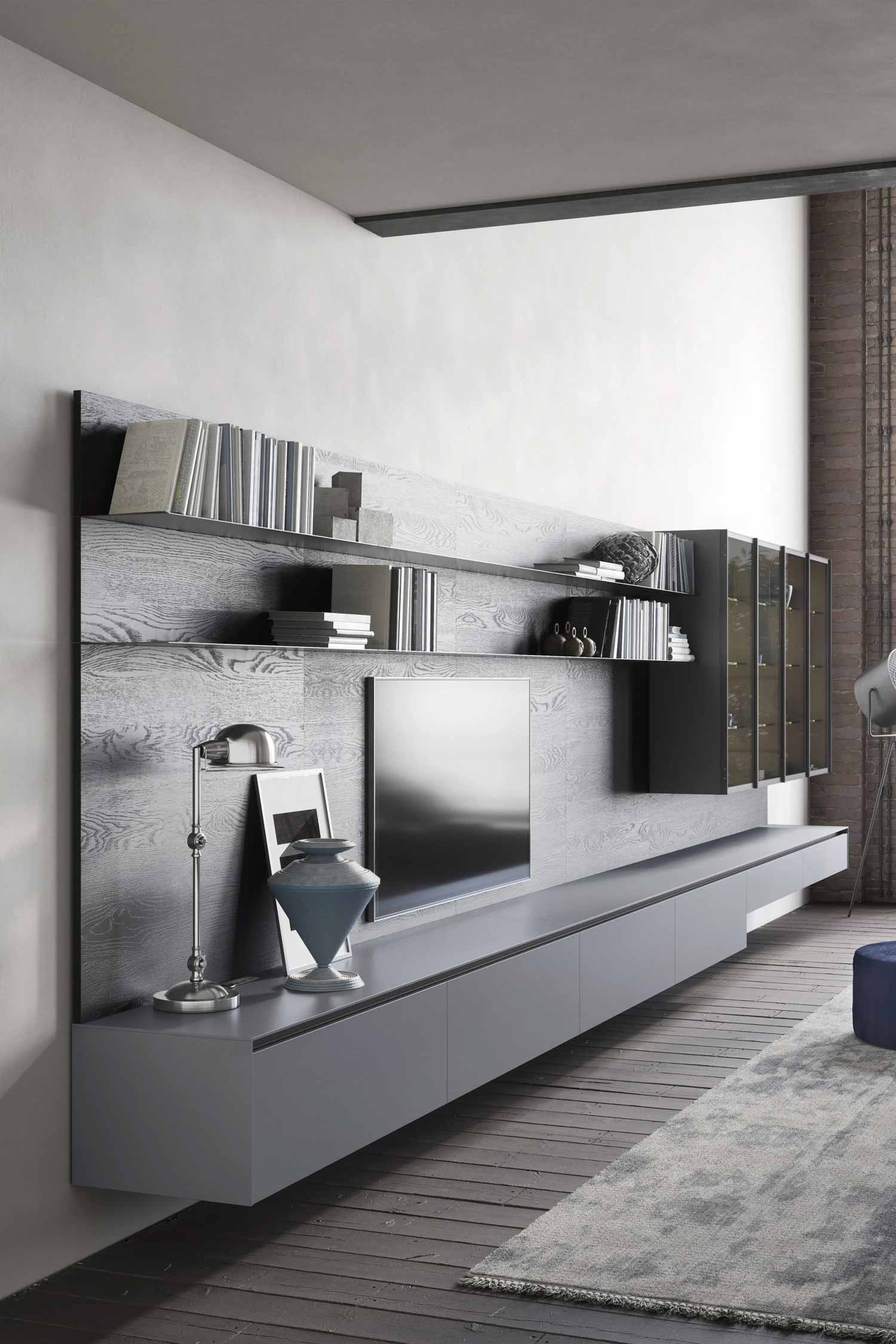 A wide-angle shot of a long, gray and dark wood-paneled media and shelving unit in a modern living room. The unit features a low, floating media console with long drawers, a mounted television in the center, and dark wood-paneled shelving above it. The shelves are filled with books, small sculptures, and decorative objects. A lit glass-front cabinet is on the far right. The floor is dark wood, and a large, textured gray rug is visible in the foreground.