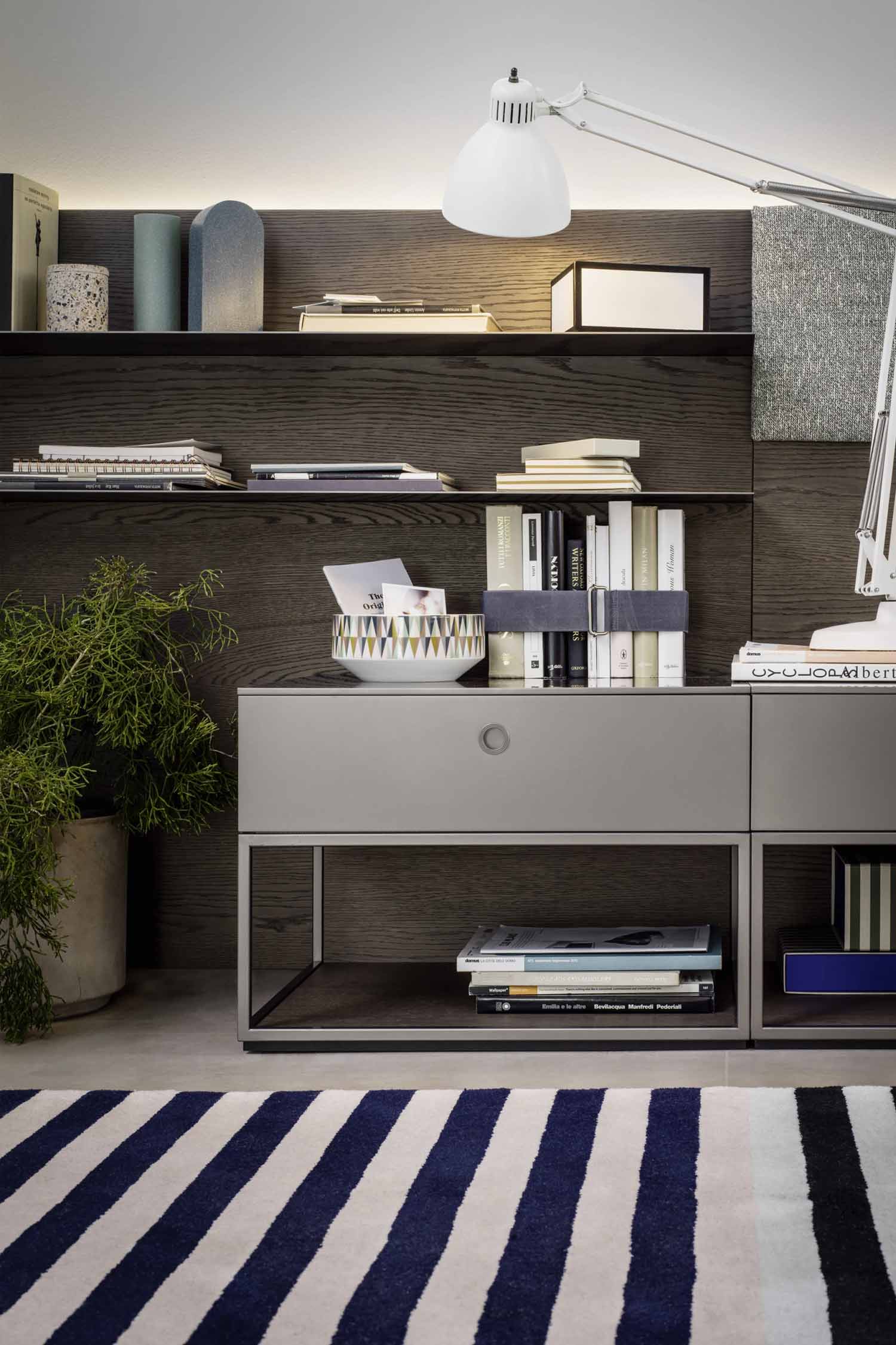 A close-up shot of a modern shelf unit with a dark wood back panel and light gray drawers. The shelves are filled with stacks of books, a decorative bowl with a colorful geometric pattern, and various art objects. A white, articulated lamp is visible on the top right. A potted plant sits on the floor to the left, and a blue and white striped rug with a spiral pattern is on the concrete floor.