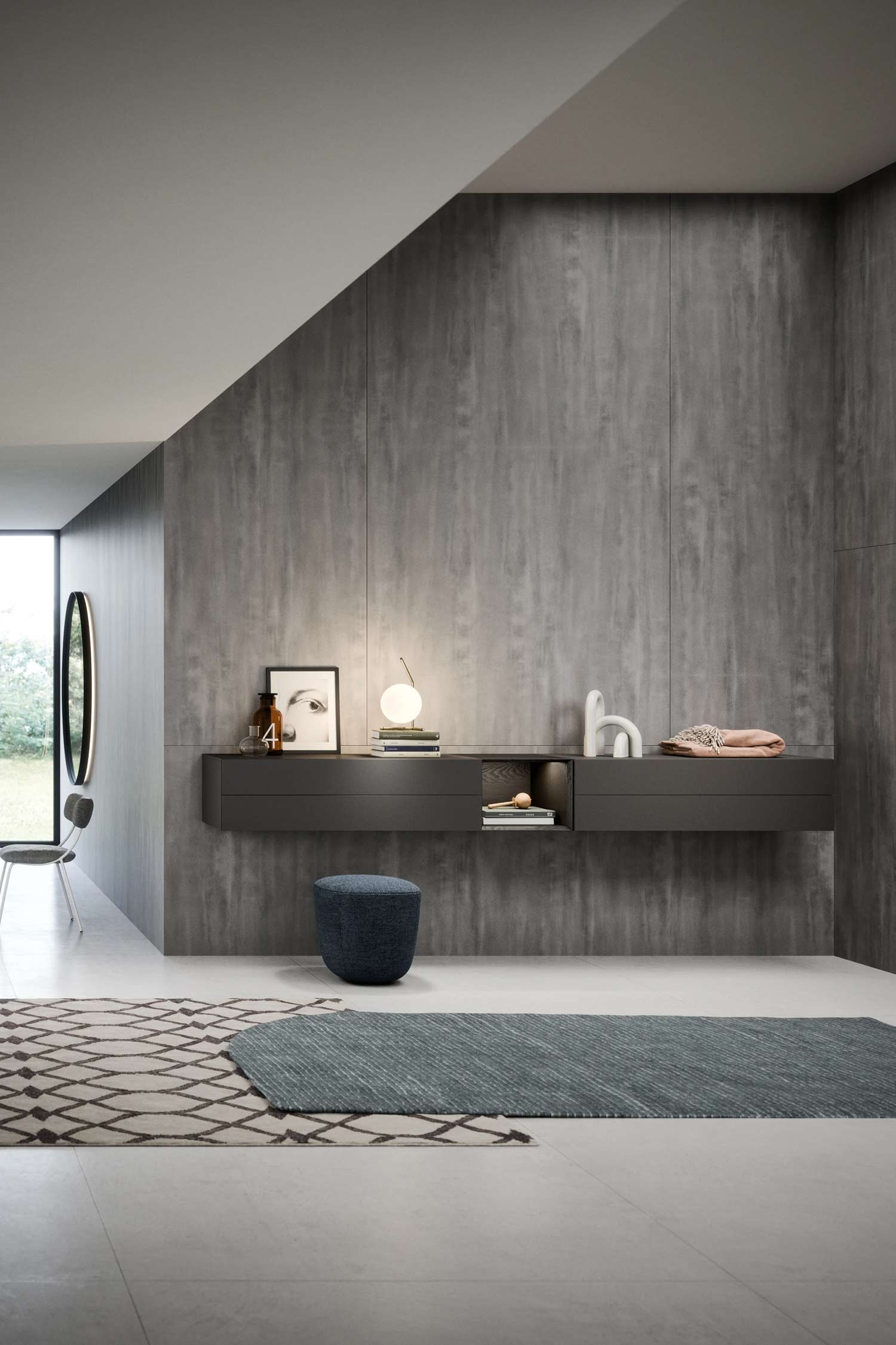 A contemporary interior space with a dramatic, floor-to-ceiling grey concrete-look accent wall. A floating dark grey console with drawers and a central cubby shelf is mounted on the wall, styled with a lamp and framed art. A small round blue ottoman sits beneath it.
