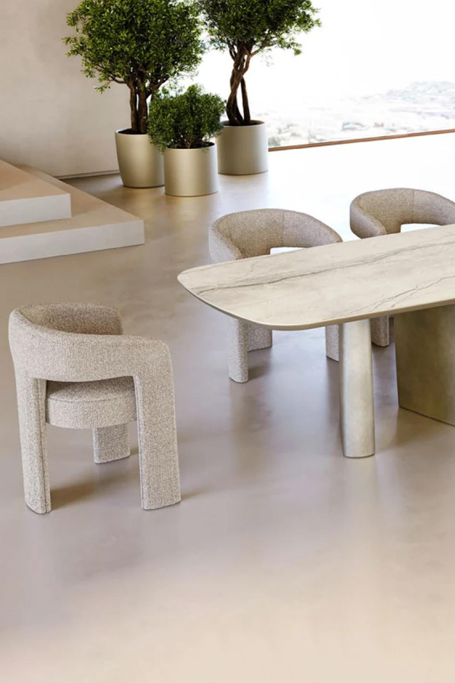 A close-up, low-angle shot showing the base of a dining table and two Egle dining chairs. The chairs are upholstered in a light, textured fabric, complementing the marble-like top and light-colored bases of the table. Large potted plants in metallic planters and an open, bright room with large windows are visible in the background.