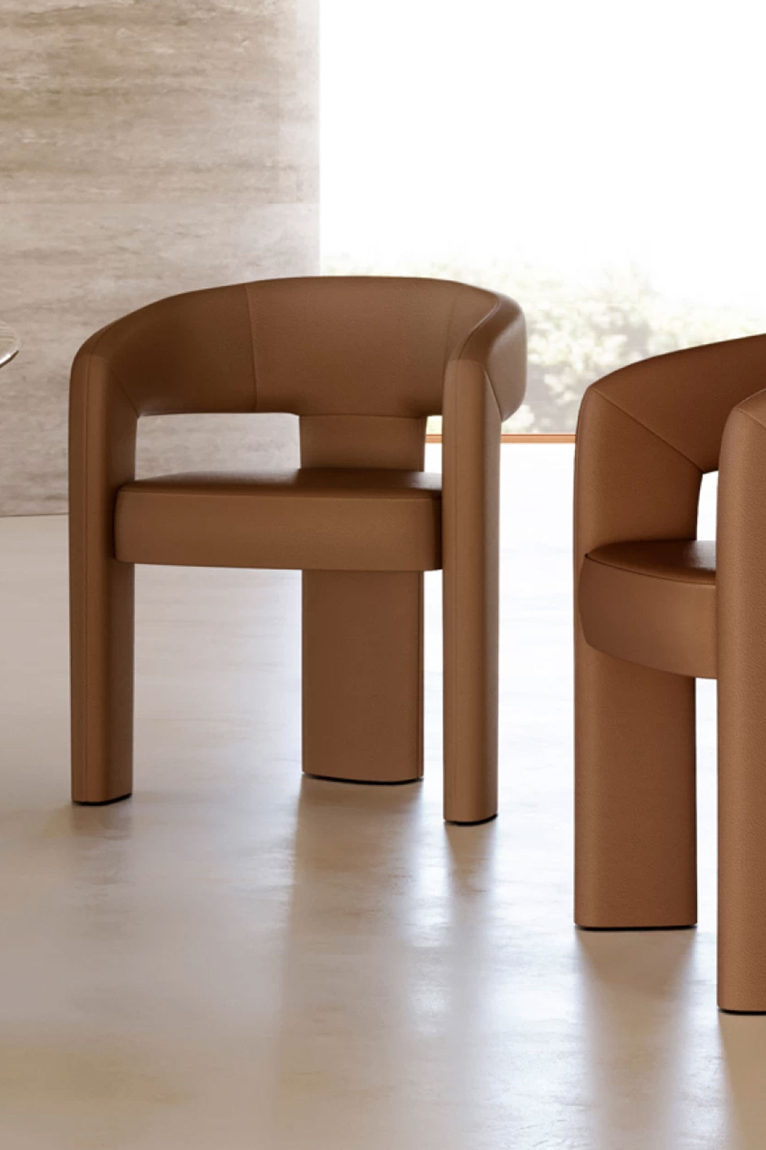 A close-up, slightly off-center shot of two Egle dining chairs upholstered in a smooth, tan-colored leather. The image focuses on the sleek material and curved shape of the chairs, with a hint of a dining table visible to the left. The background shows a light-filled modern room with large windows, emphasizing the chairs as a focal point.
