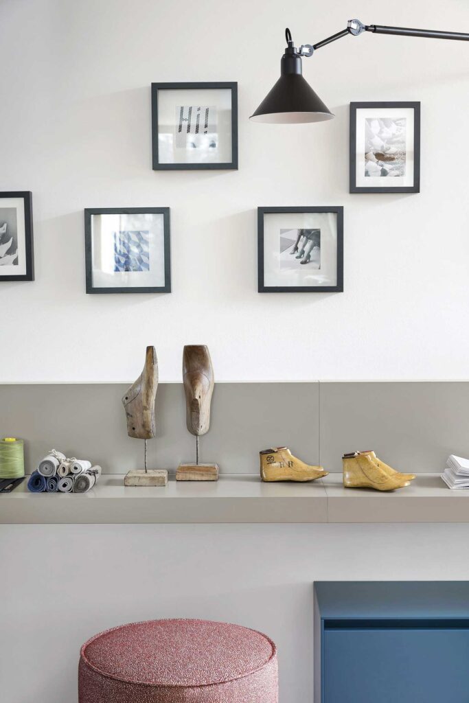 A close-up of a modern, minimal, grey console table. The table is mounted on a white wall, and on top of it are several decorative objects, including two shoe lasts on metal stands, and two more modern sculptures of shoes. A collection of small rolls of fabric and a spool of green thread are visible on the far left. A round, pink pouf is visible in the bottom left of the frame. Above the table, an arrangement of five black-framed pieces of art are hung on the wall. A black, articulated wall lamp is mounted at the top of the frame.