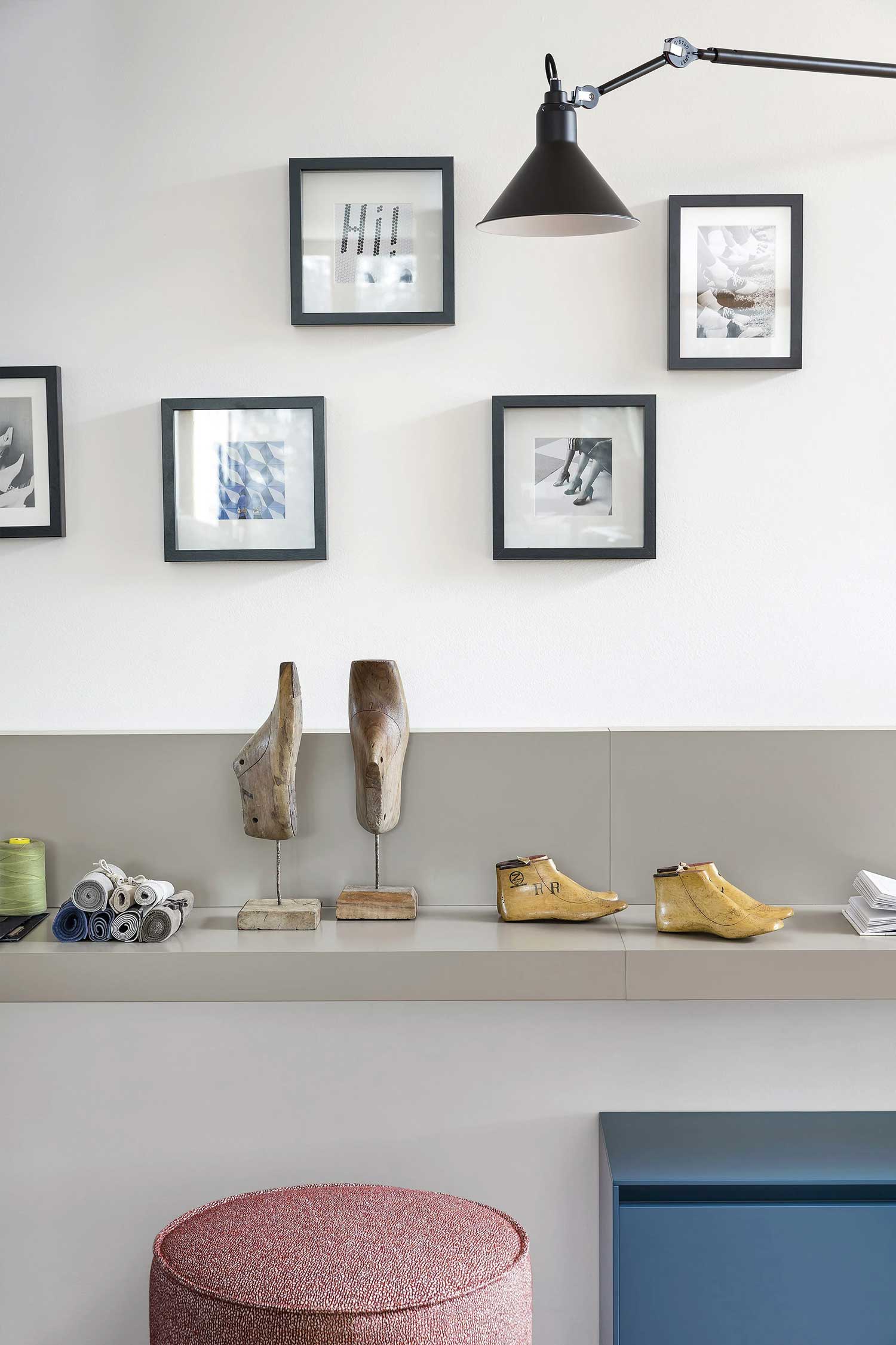 A close-up of a modern, minimal, grey console table. The table is mounted on a white wall, and on top of it are several decorative objects, including two shoe lasts on metal stands, and two more modern sculptures of shoes. A collection of small rolls of fabric and a spool of green thread are visible on the far left. A round, pink pouf is visible in the bottom left of the frame. Above the table, an arrangement of five black-framed pieces of art are hung on the wall. A black, articulated wall lamp is mounted at the top of the frame.