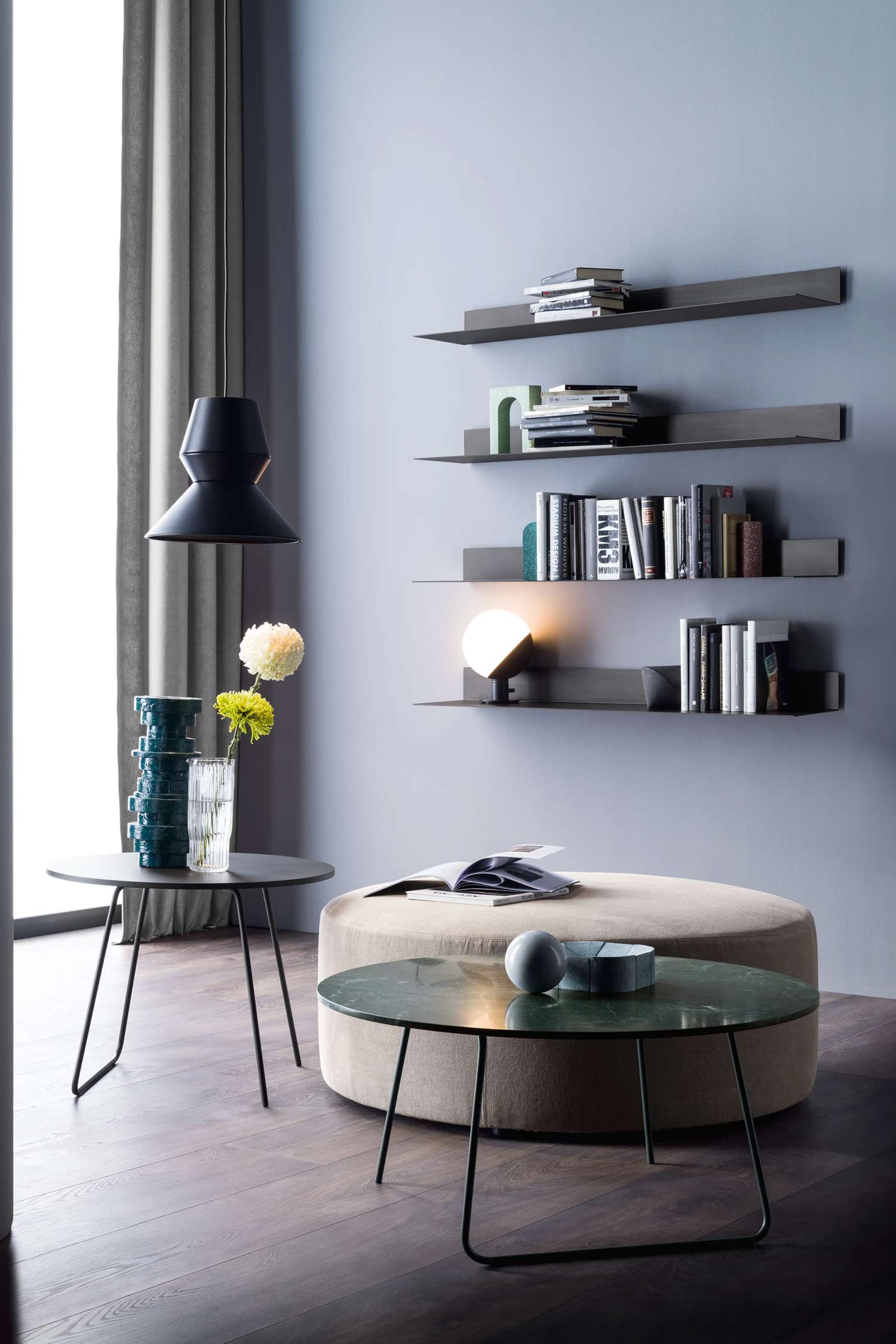 A living space features a large, round ottoman with a beige fabric top, positioned on a dark wood floor. A round coffee table with a green marble top and black metal legs sits in front of the ottoman, holding a gray decorative sphere and a box. To the left, a smaller, round side table with a black top holds a clear vase with yellow and white flowers. In the background, three dark floating shelves on a light blue-gray wall are decorated with books and modern art.