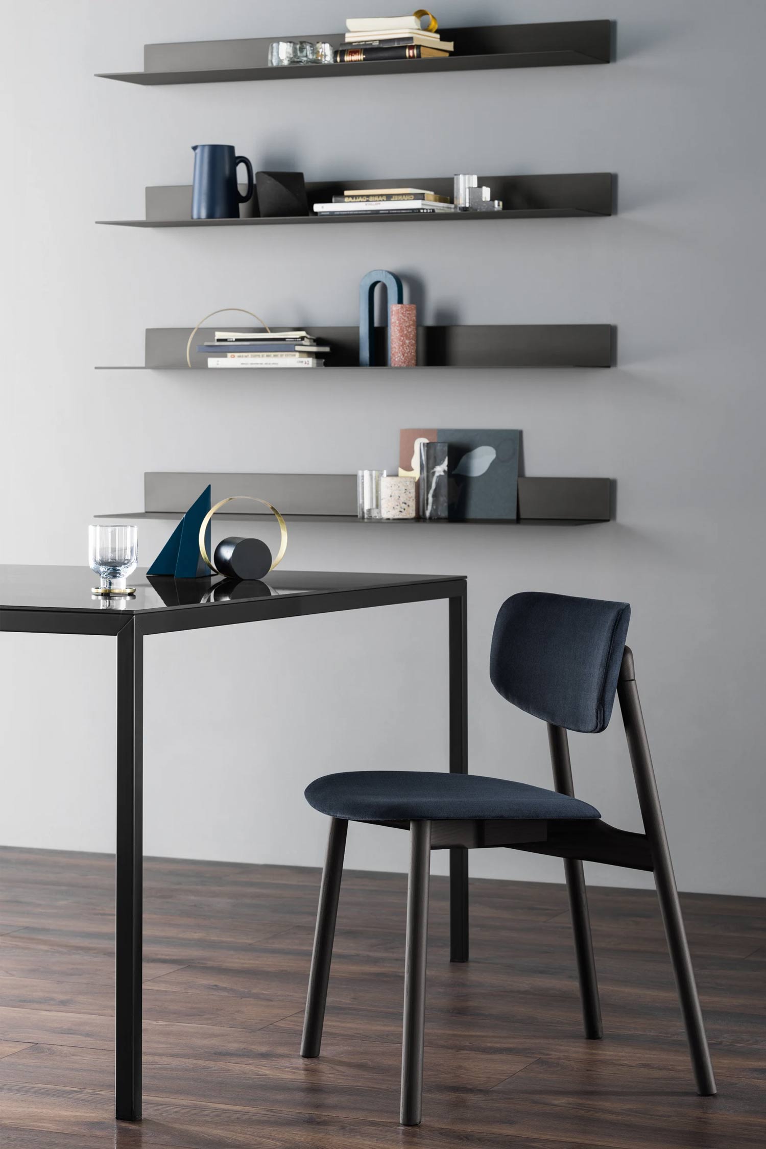A living space features a large, round ottoman with a beige fabric top, positioned on a dark wood floor. A round coffee table with a green marble top and black metal legs sits in front of the ottoman, holding a gray decorative sphere and a box. To the left, a smaller, round side table with a black top holds a clear vase with yellow and white flowers. In the background, three dark floating shelves on a light blue-gray wall are decorated with books and modern art.