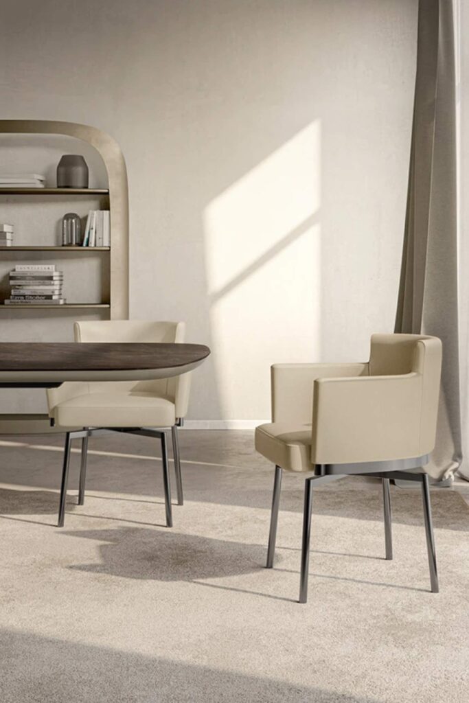 Two cream-colored, contemporary dining chairs with thin metal legs are shown in a modern dining room with a large window and a dark wood table. A curved bookshelf is visible in the background.