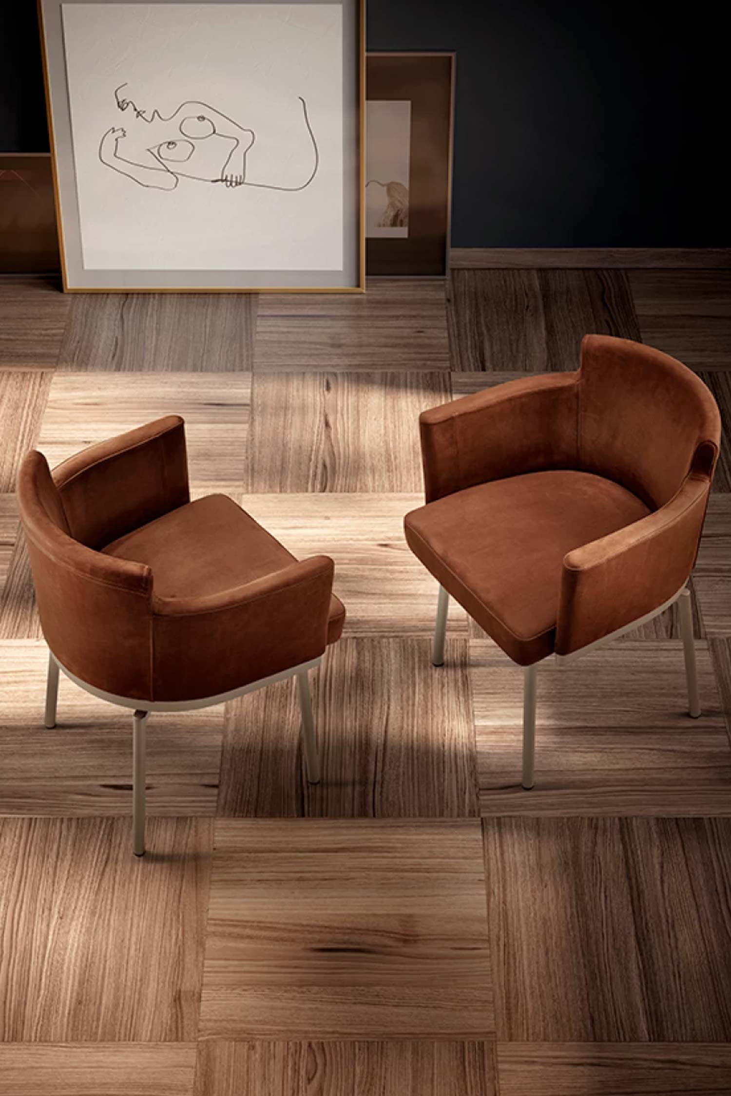 Two brown, barrel-style chairs with white legs are placed on a checkered wood floor. A framed art piece hangs on the dark wall behind them.