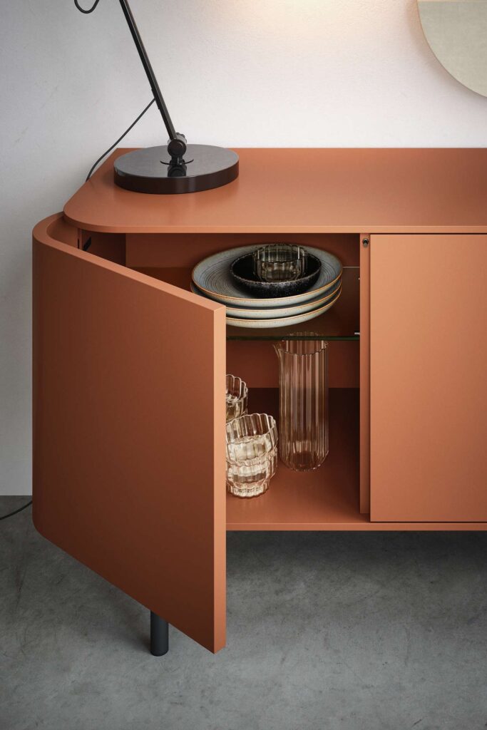 A close-up of a modern, terracotta-colored sideboard with one door open. The interior shelf holds a stack of dark-rimmed plates and a striped glass bowl. The bottom section holds a ribbed glass carafe and two textured glasses. A black desk lamp is on top of the sideboard.